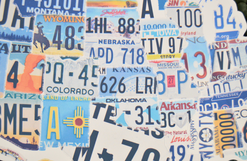 A square art print or canvas featuring a dense collage of colorful vintage license plates from various states, including Nebraska, Arizona, Oklahoma, Texas, and Wyoming, arranged in a textured pattern.