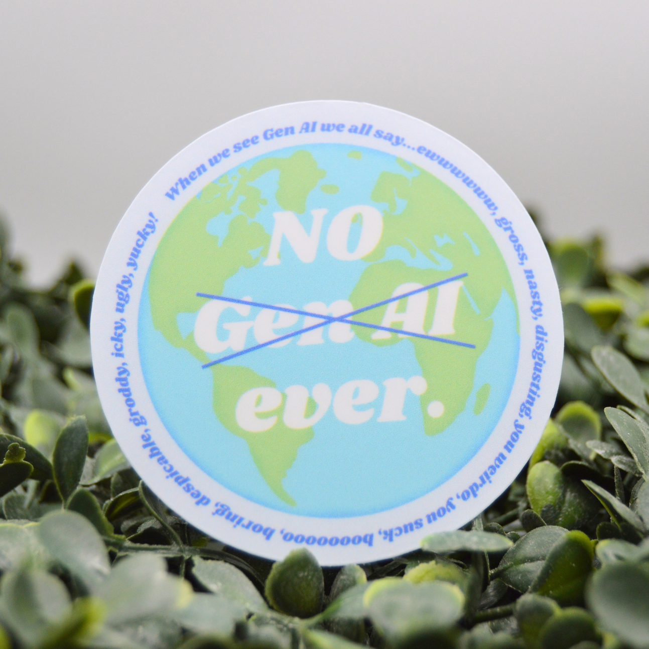 A circular sticker featuring a light blue and green globe design with the text 'NO Gen Al ever.' crossed out with a blue diagonal line, placed on a bed of green leaves.