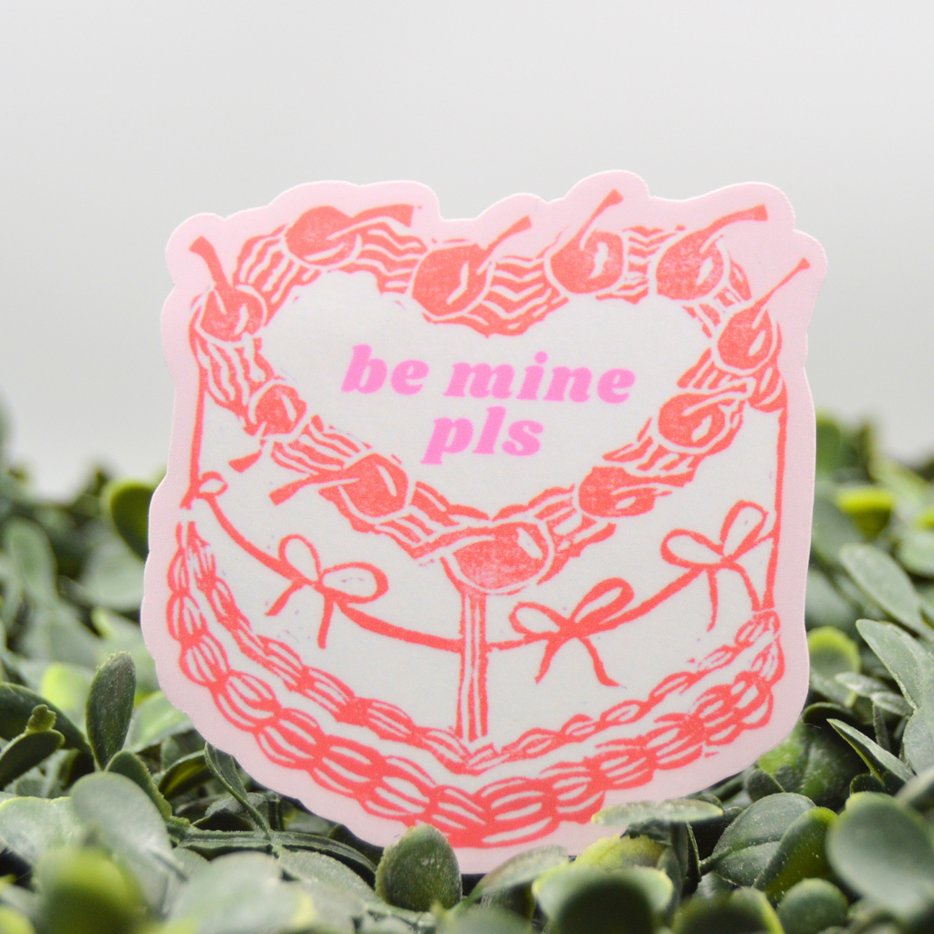 A die-cut sticker featuring a pink heart-shaped wreath with cherries and the text 'be mine pls' in pink, shown standing upright on green foliage against a light gray background.