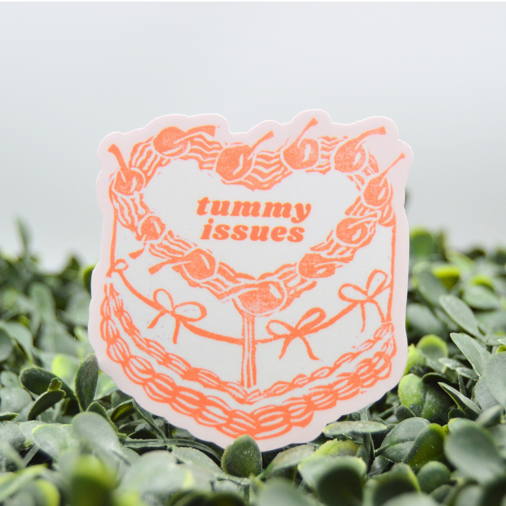 A die-cut sticker with a white background and orange graphic of a decorated cake with the text 'tummy issues' in the center, placed on green foliage against a light gray background.