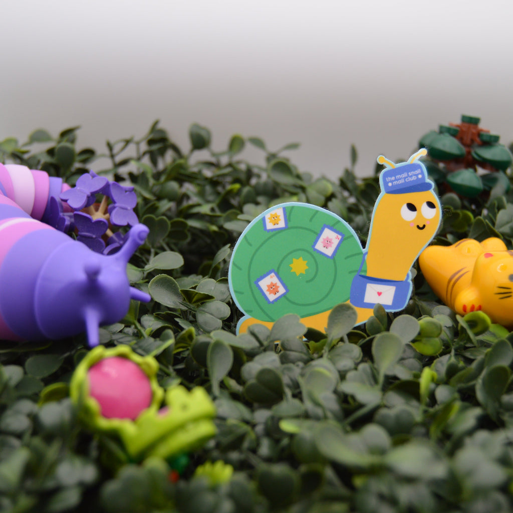 A set of colorful children's toy figures arranged on green artificial foliage. The set includes a purple and pink striped caterpillar, a green snail with a blue hat, a yellow cat, and small flower pieces.