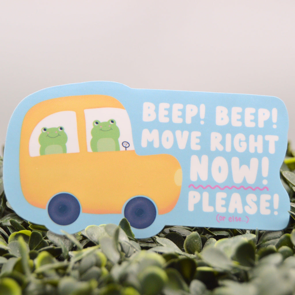 A die-cut decorative sign featuring a yellow cartoon car with two green frogs and the text 'BEEP! BEEP! MOVE RIGHT NOW! PLEASE!' on a light blue background, placed on green foliage.