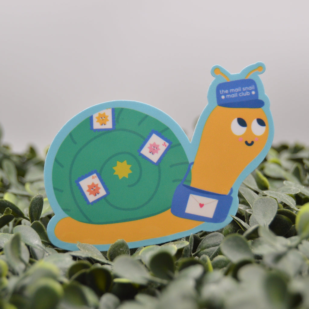 A die-cut paper snail sign with a green shell and orange body, wearing a blue hat and belt, standing on green foliage against a light gray background.