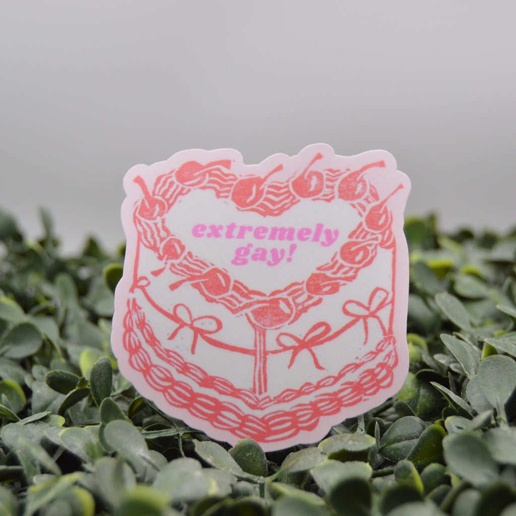 A die-cut sticker featuring a pink and white illustrated cake with the text 'extremely gay!' in pink, displayed on a bed of green artificial foliage against a soft gray background.