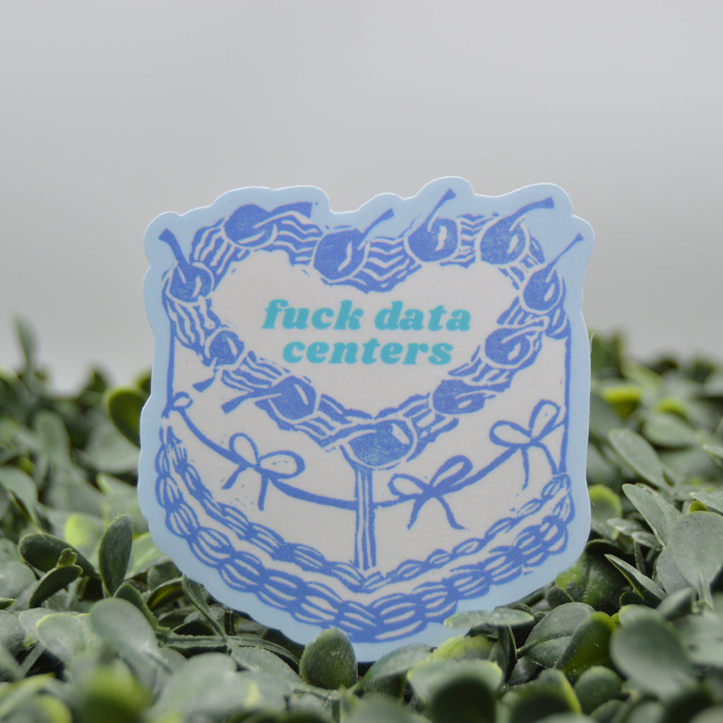 A die-cut sticker featuring a blue cake illustration with the text 'fuck data centers' in teal, displayed on a bed of green foliage against a soft gray background.