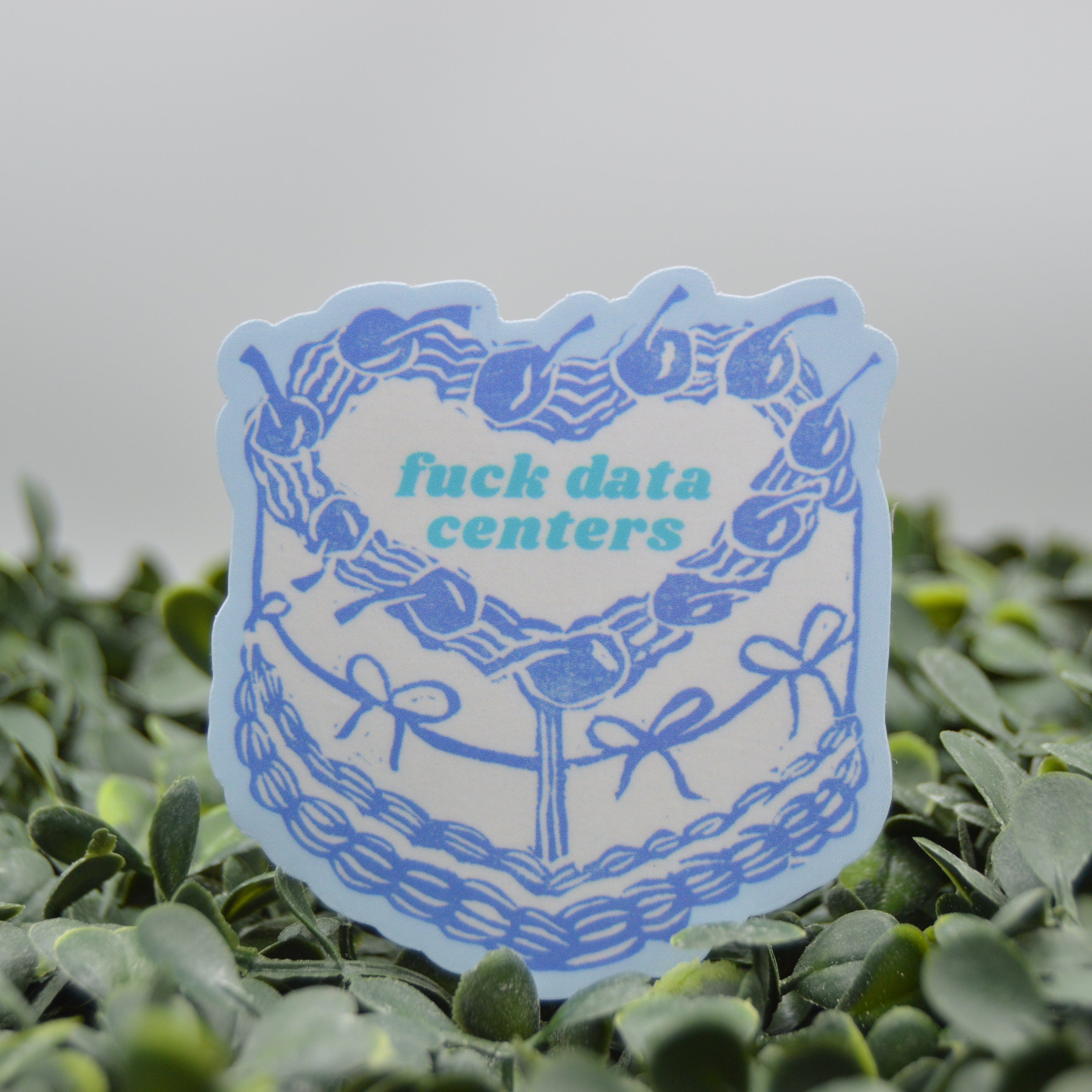A die-cut sticker featuring a blue cake illustration with the text 'fuck data centers' in teal, displayed on a bed of green foliage against a soft gray background.