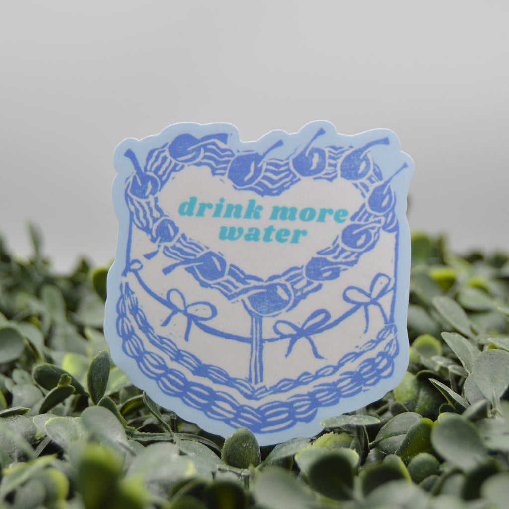 A die-cut sticker featuring a blue cake illustration with cherries and the text 'drink more water' in teal, displayed on a bed of green leaves against a soft gray background.