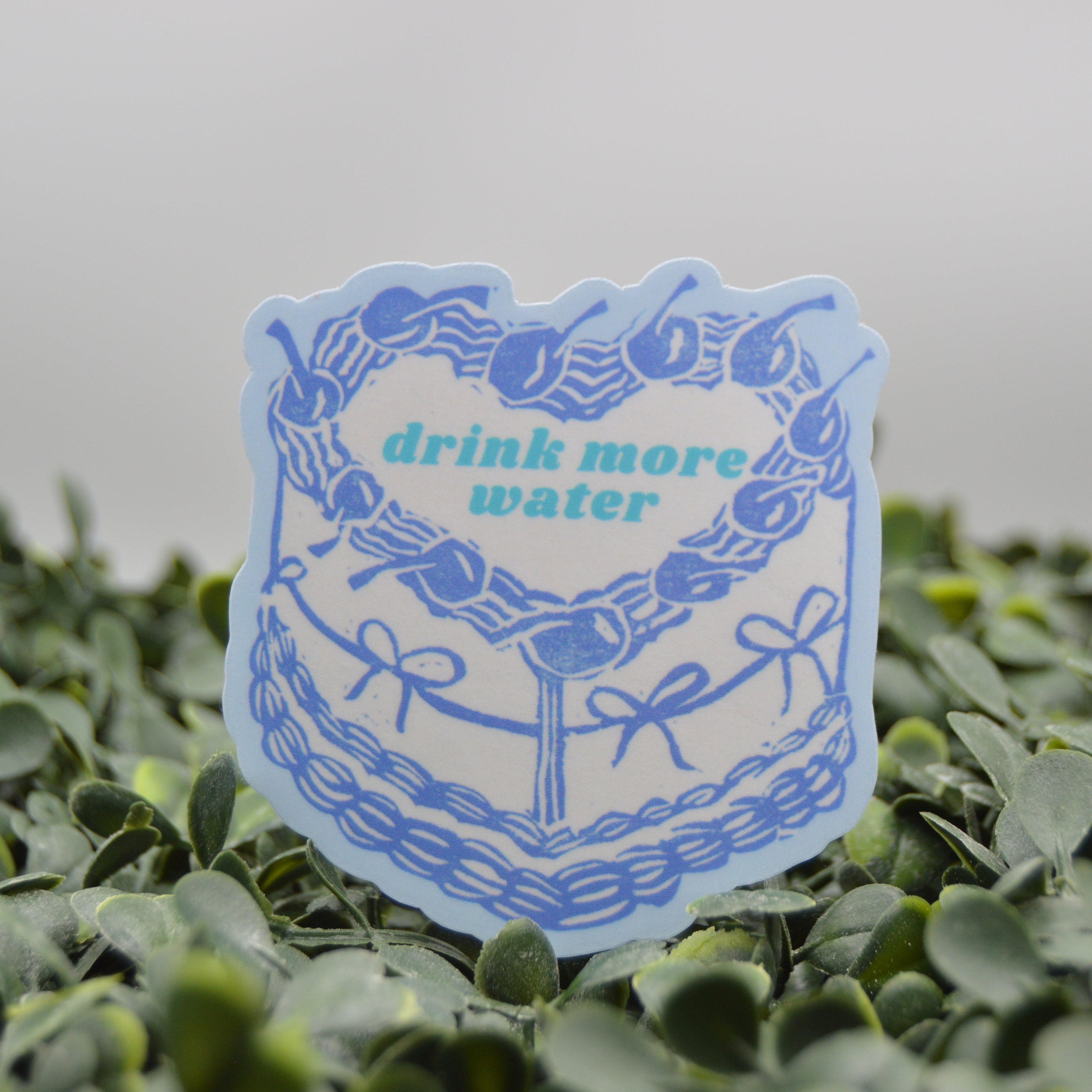 A die-cut sticker featuring a blue cake illustration with cherries and the text 'drink more water' in teal, displayed on a bed of green leaves against a soft gray background.