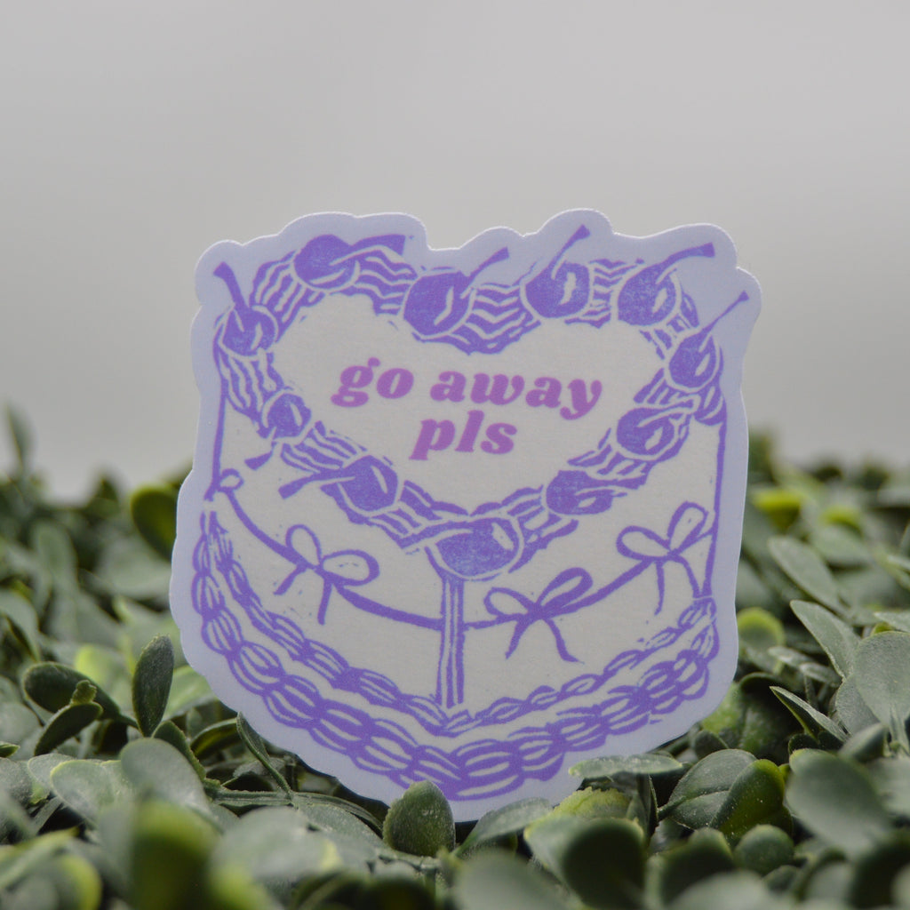 A die-cut sticker featuring a purple heart-shaped wreath with cherries and the text 'go away pls' in purple, shown standing upright in green foliage against a soft gray background.