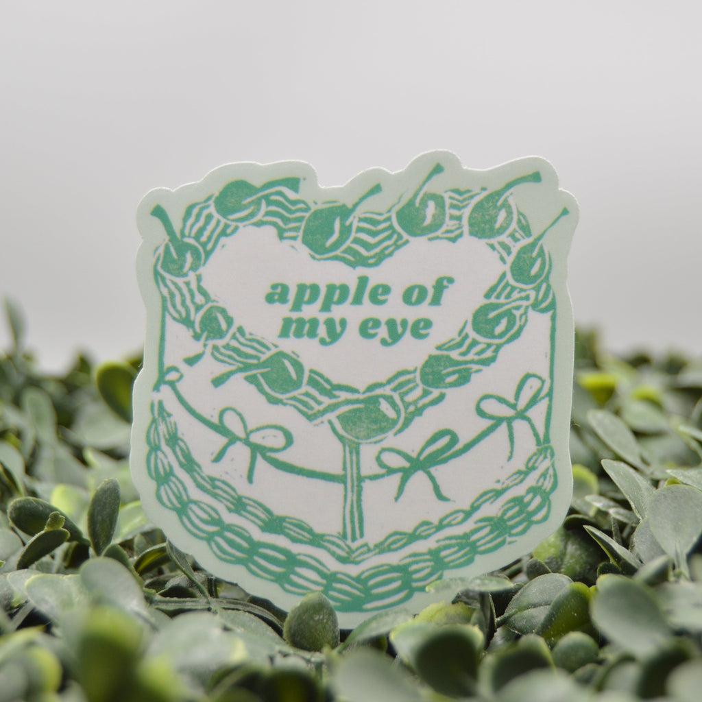 A die-cut sticker featuring a green cake design with the text 'apple of my eye' in green, displayed on a bed of small green leaves against a soft gray background.