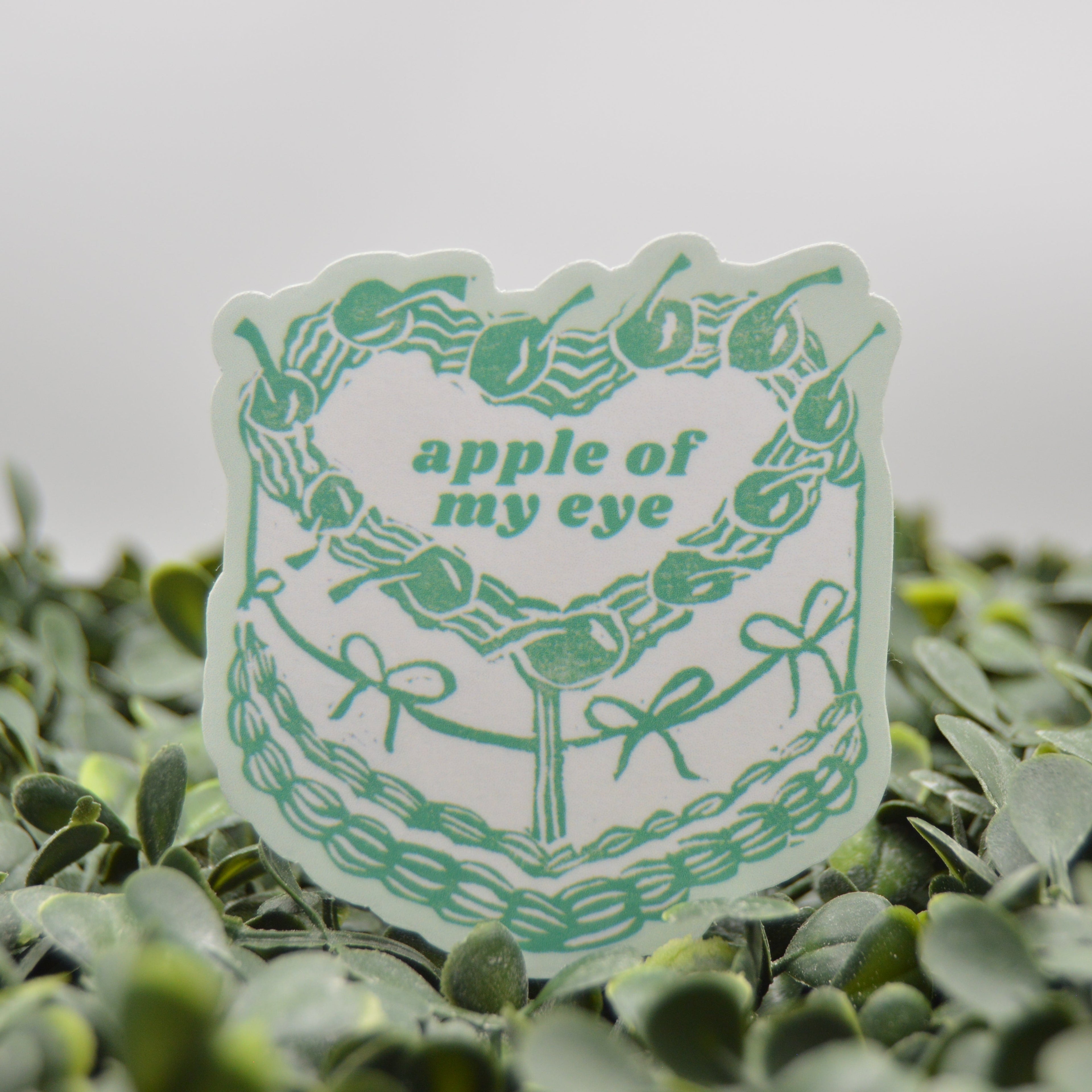 A die-cut sticker featuring a green cake design with the text 'apple of my eye' in green, displayed on a bed of small green leaves against a soft gray background.
