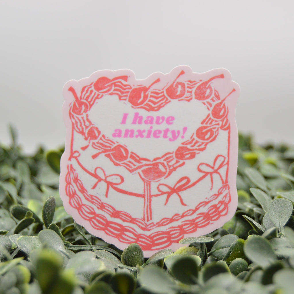 A die-cut sticker featuring a pink cherry wreath with the text 'I have anxiety!' in pink, placed on green foliage against a soft gray background.