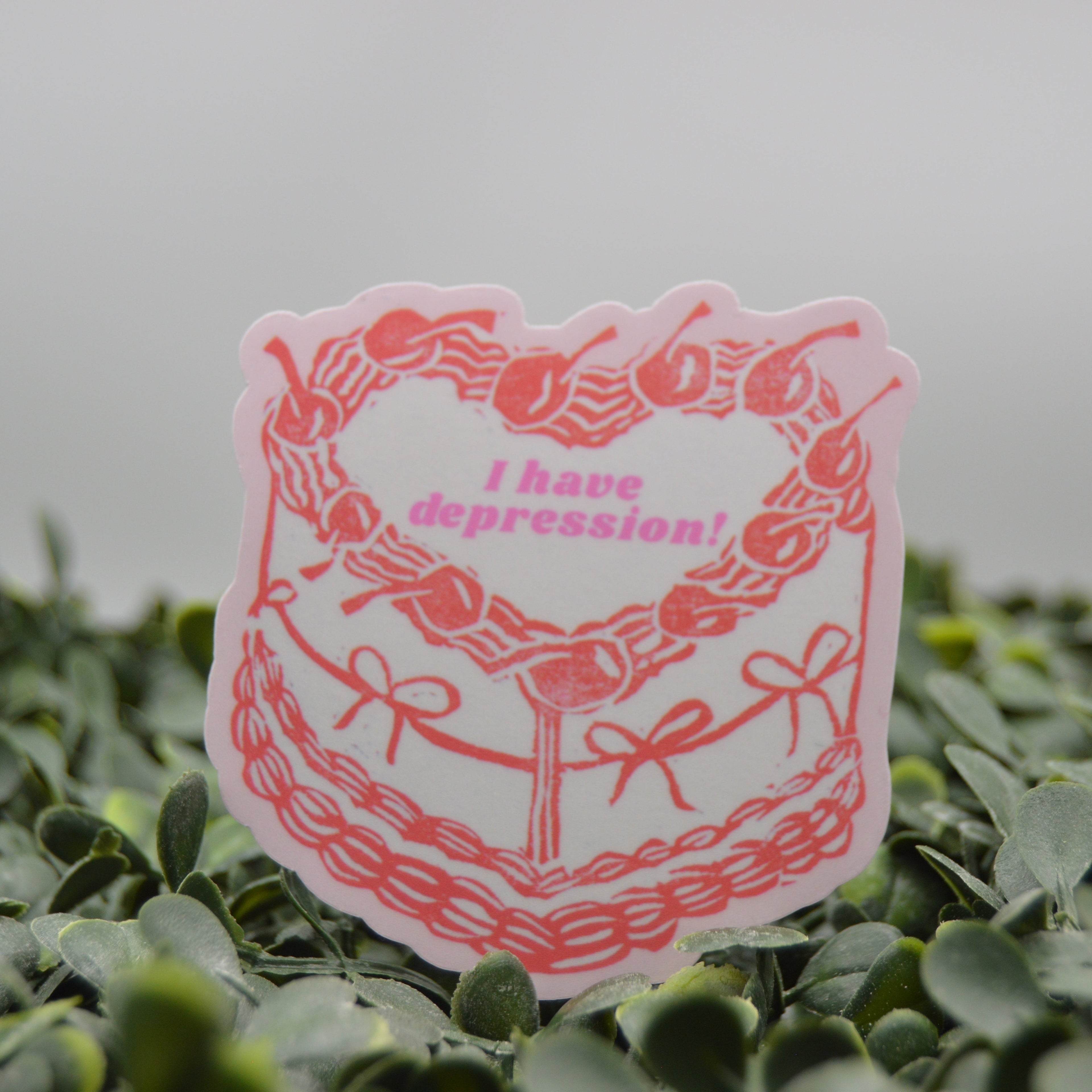 A die-cut sticker featuring a pink cherry wreath and the text 'I have depression!' in pink, displayed on a bed of green leaves against a soft gray background.