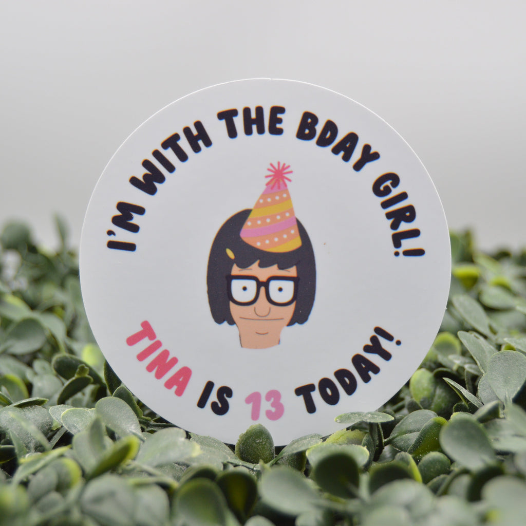 A round white sticker with a cartoon character and the text 'I'M WITH THE BDAY GIRL! TINA IS 13 TODAY!' placed on green foliage against a light gray background.