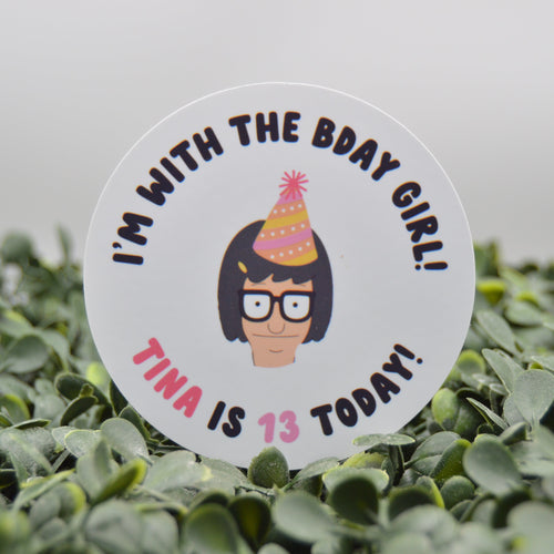 A round white sticker with a cartoon character and the text 'I'M WITH THE BDAY GIRL! TINA IS 13 TODAY!' placed on green foliage against a light gray background.