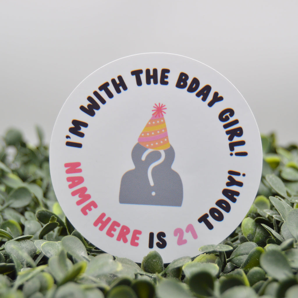 A round white sticker with a 21st birthday theme, featuring a gray silhouette wearing a pink and yellow party hat, surrounded by the text 'I'M WITH THE BDAY GIRL! NAME HERE IS 21 TODAY!' in black and pink lettering. The sticker is displayed upright in a bed of small green leaves against a soft, out-of-focus light gray background.