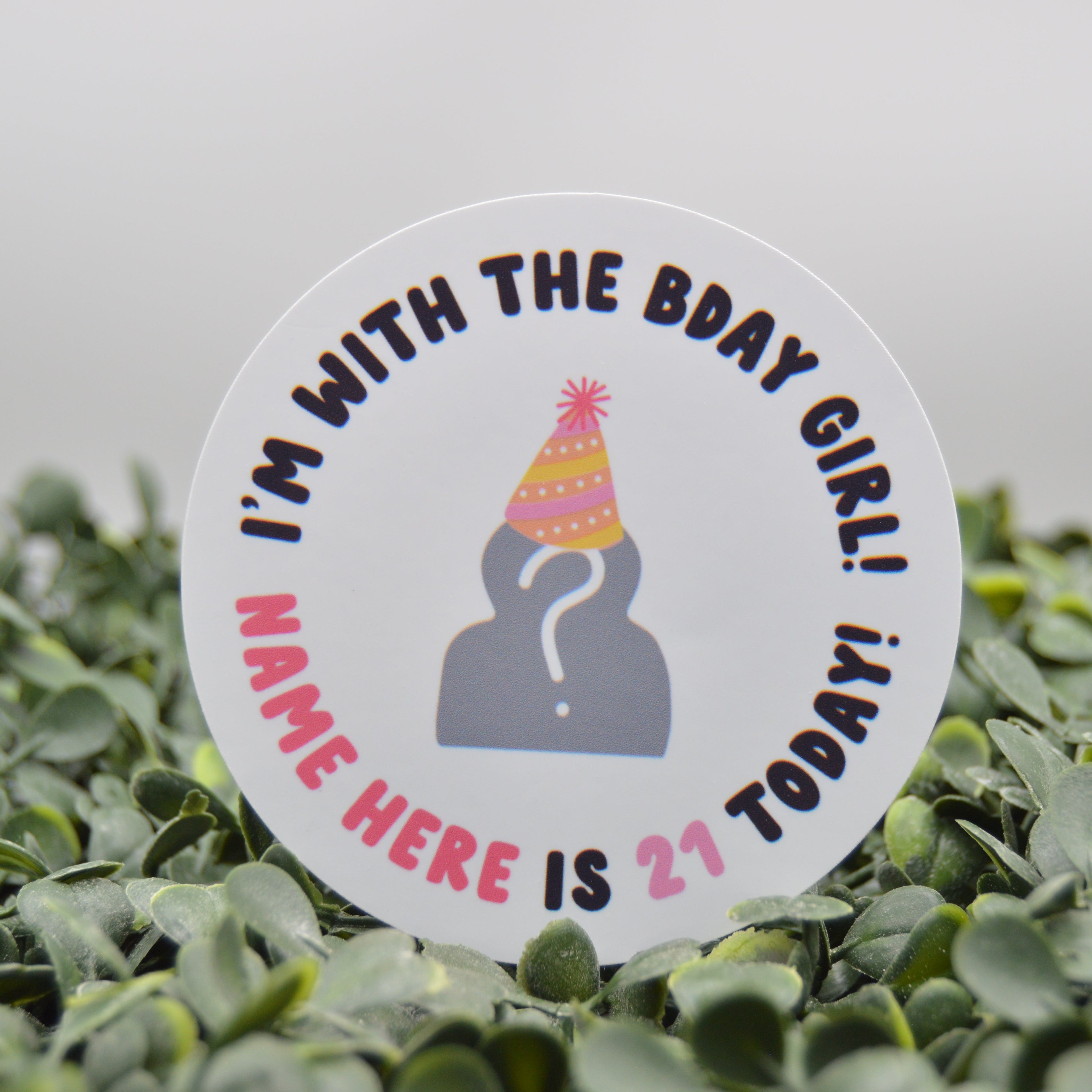 A round white sticker with a 21st birthday theme, featuring a gray silhouette wearing a pink and yellow party hat, surrounded by the text 'I'M WITH THE BDAY GIRL! NAME HERE IS 21 TODAY!' in black and pink lettering. The sticker is displayed upright in a bed of small green leaves against a soft, out-of-focus light gray background.