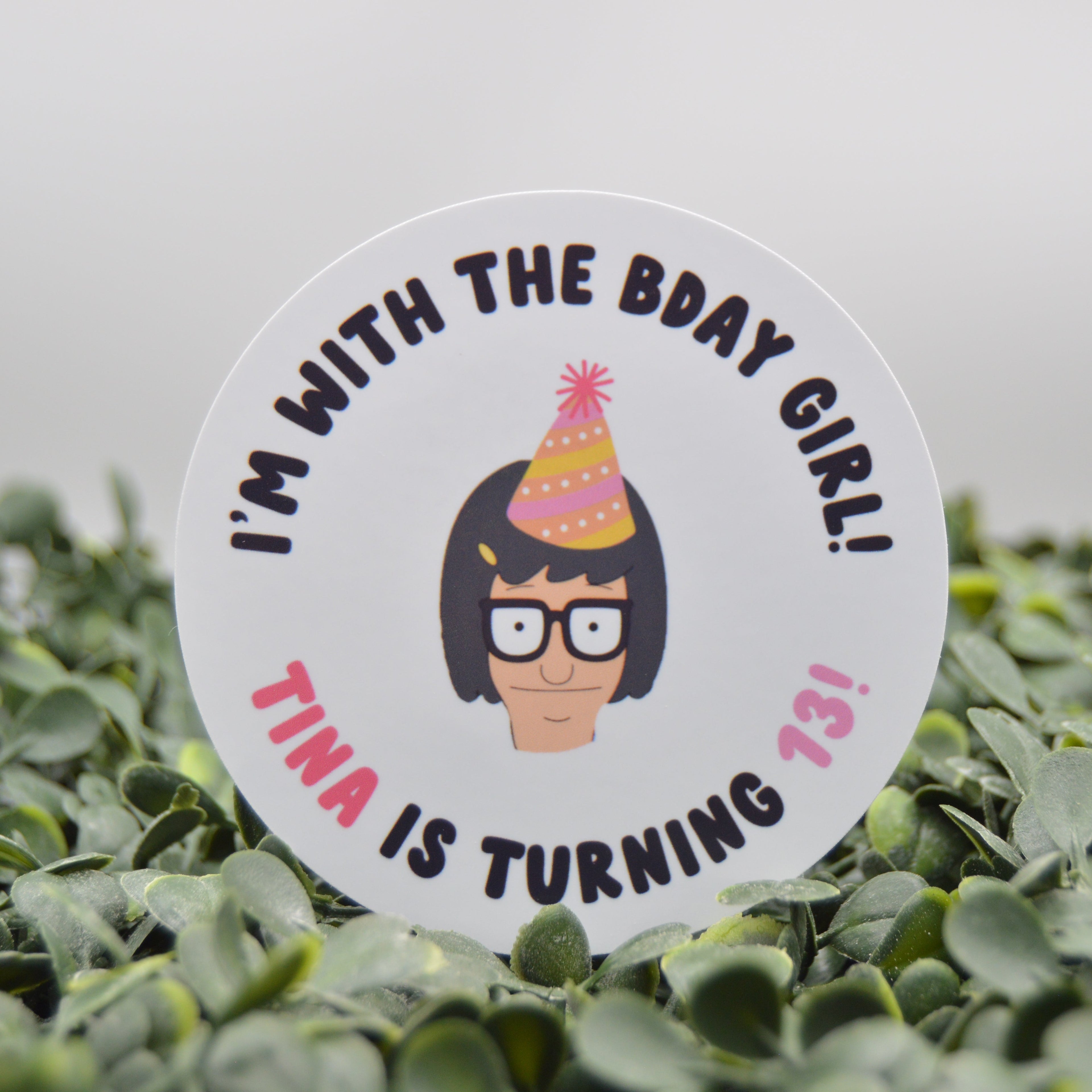 A round white sticker featuring an illustration of Tina from BoJack Bunny wearing a party hat, surrounded by the text 'I'M WITH THE BDAY GIRL! TINA IS TURNING 13!' in black and pink lettering. The sticker is displayed upright on a bed of green leaves against a soft gray background.