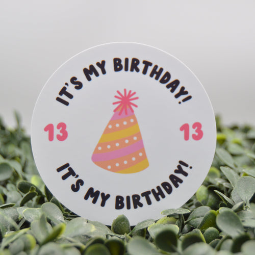 A round white birthday candle topper sticker with the text 'IT'S MY BIRTHDAY!' and a colorful party hat illustration, displayed on green foliage against a soft gray background.
