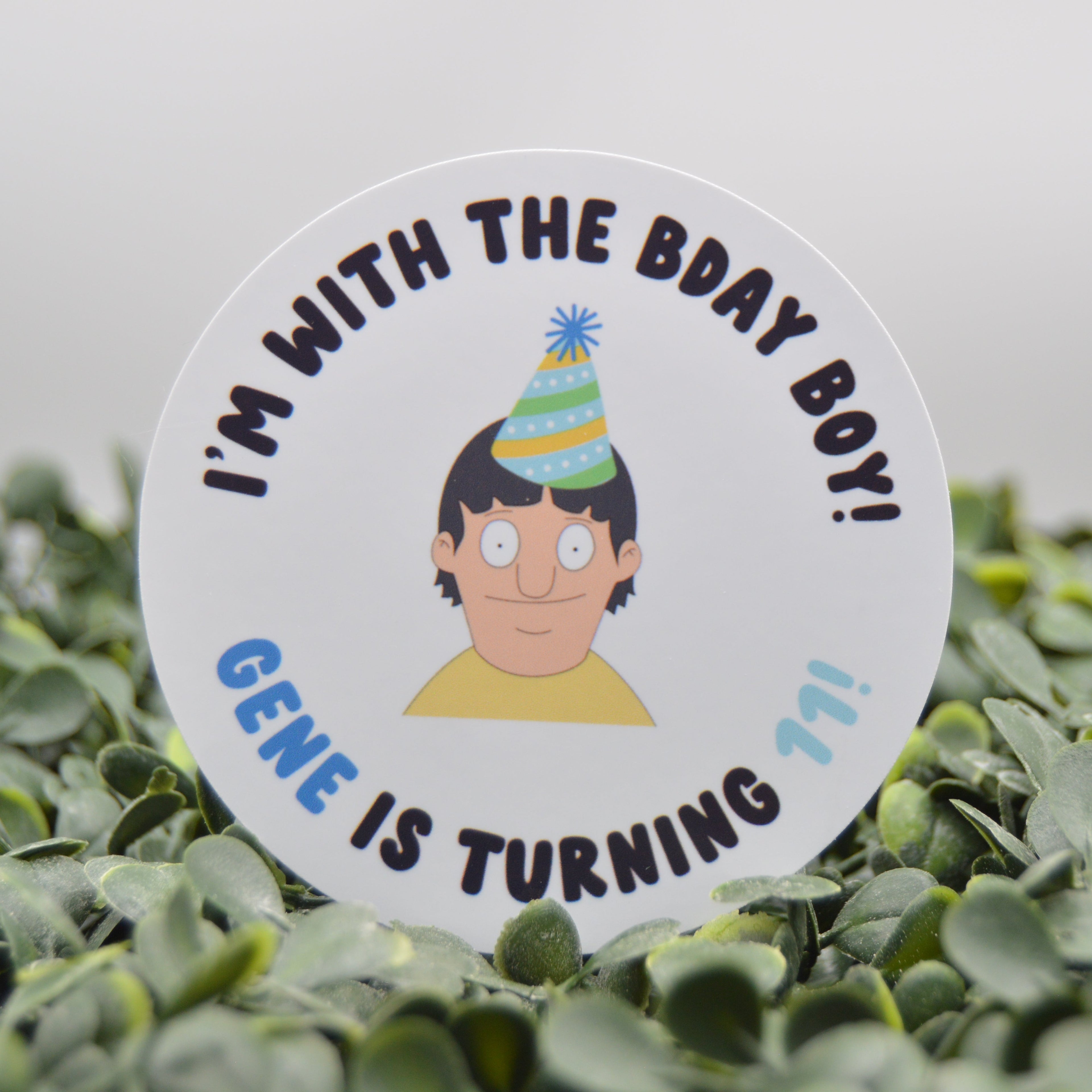 A round white party favor tag or sticker featuring a cartoon illustration of Boing Boing wearing a striped party hat, with the text 'I'M WITH THE BDAY BOY! GENE IS TURNING 11!!' in blue and black lettering. The tag is displayed upright among green leaves against a soft gray background.