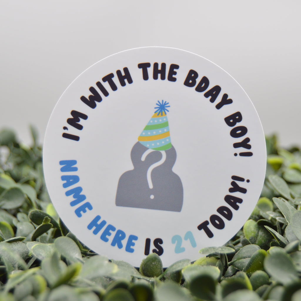 A round white sticker with a party hat illustration and the text 'I'M WITH THE BDAY BOY! NAME HERE IS 21 TODAY!' displayed upright in green foliage against a soft gray background.