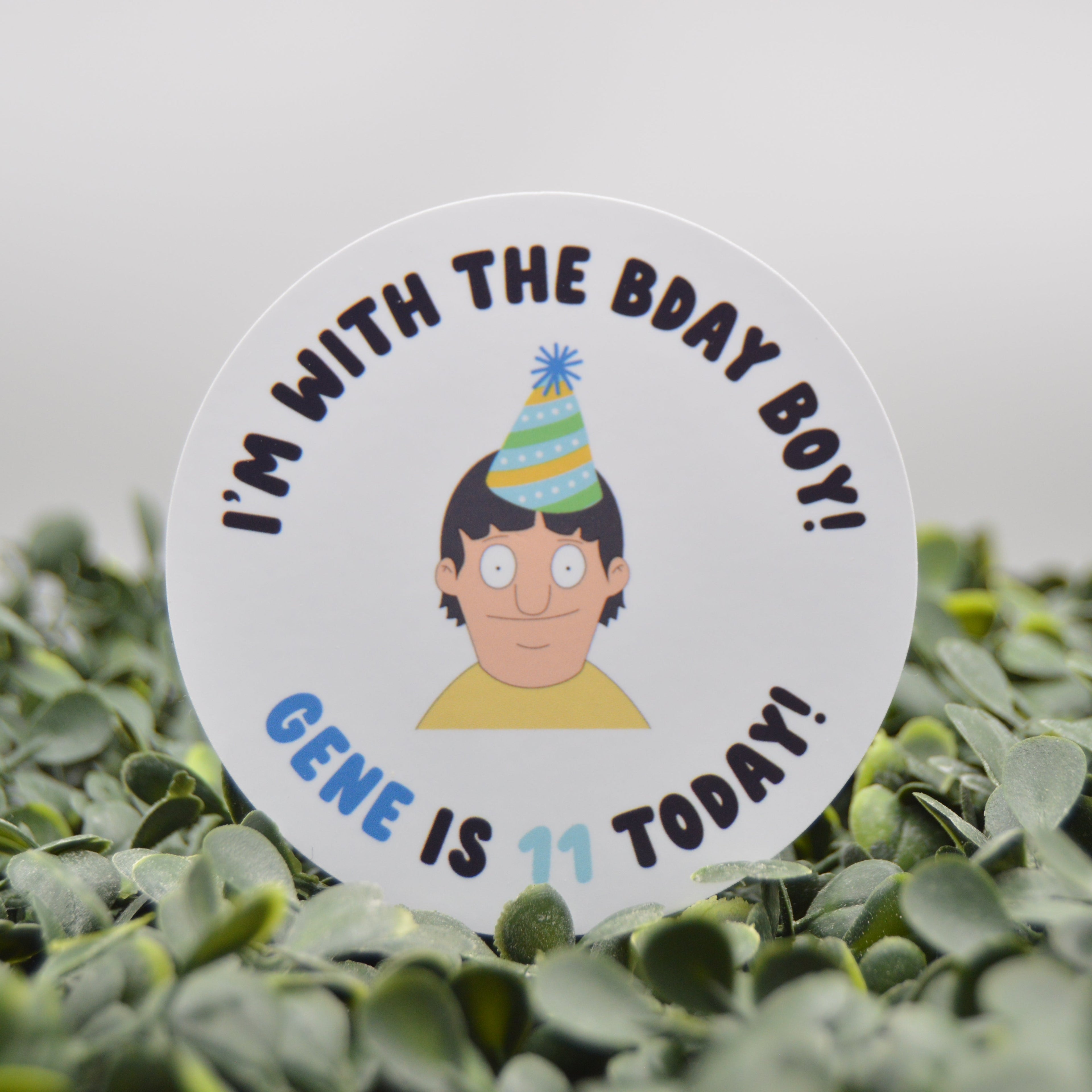 A round white sticker featuring a cartoon character wearing a striped party hat, surrounded by the text 'I'M WITH THE BDAY BOY! GENE IS 11 TODAY!' in blue and black lettering. The sticker is placed on a bed of green leaves against a soft gray background.