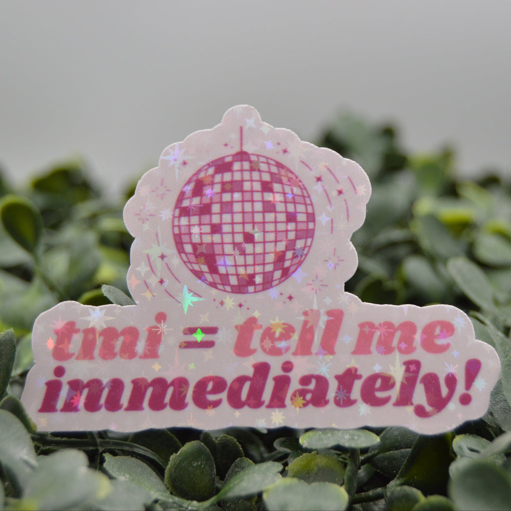 A pink and white die-cut sticker featuring a disco ball graphic and the text 'tmi = tell me immediately!' in pink, displayed on a bed of green foliage against a soft gray background.