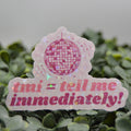 A pink and white die-cut sticker featuring a disco ball graphic and the text 'tmi = tell me immediately!' in pink, displayed on a bed of green foliage against a soft gray background.