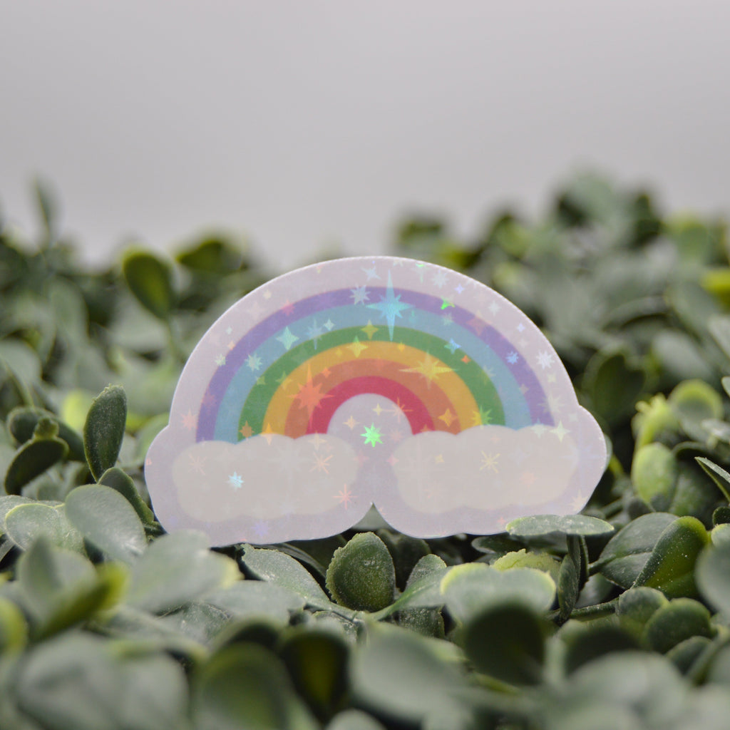 A small holographic rainbow and cloud sticker or charm placed on dense green foliage against a soft gray background.