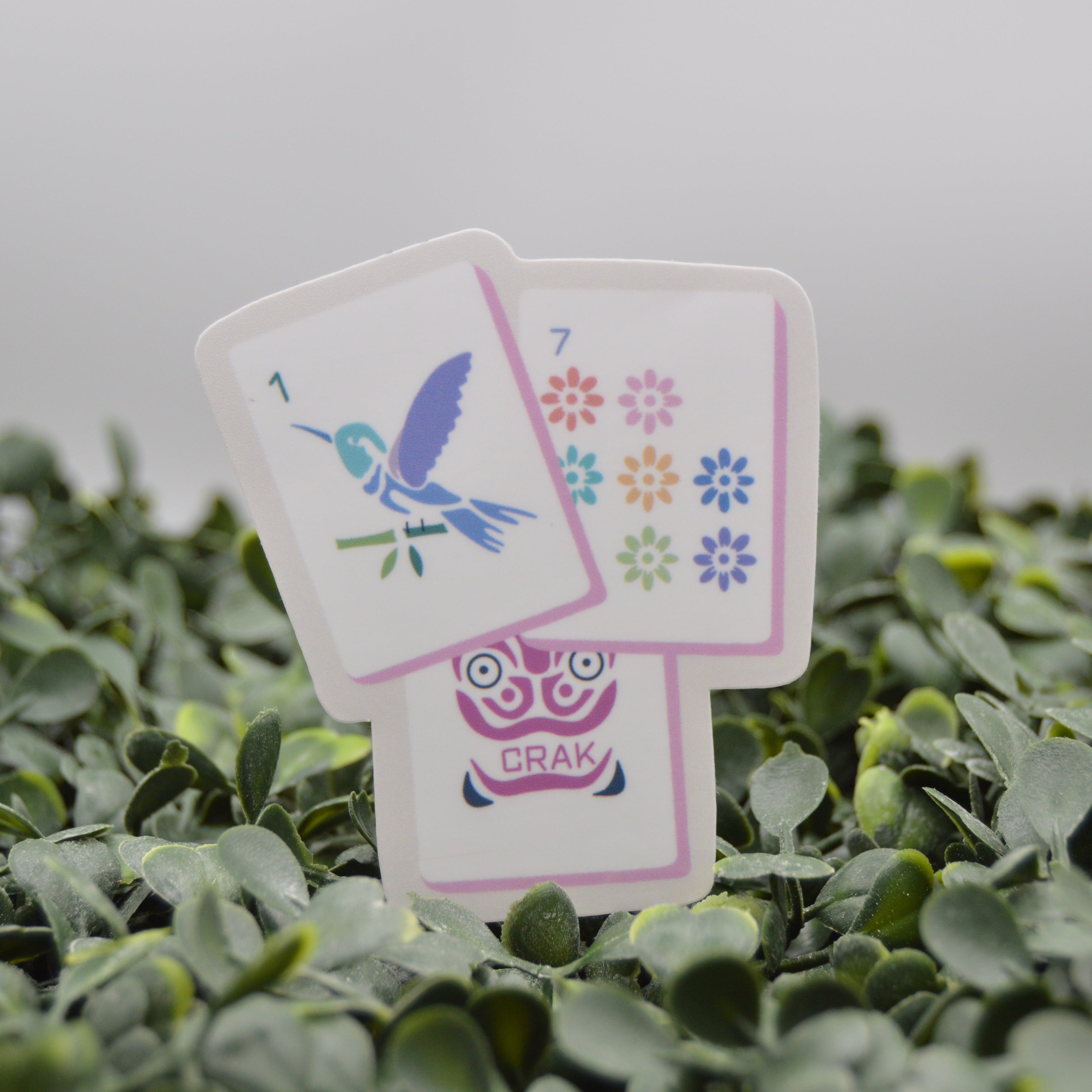 Three illustrated cards standing upright in dense green foliage. The front card shows a blue hummingbird and the number 1, the middle card displays colorful flower symbols, and the back card shows a pink owl with the word CRAK.