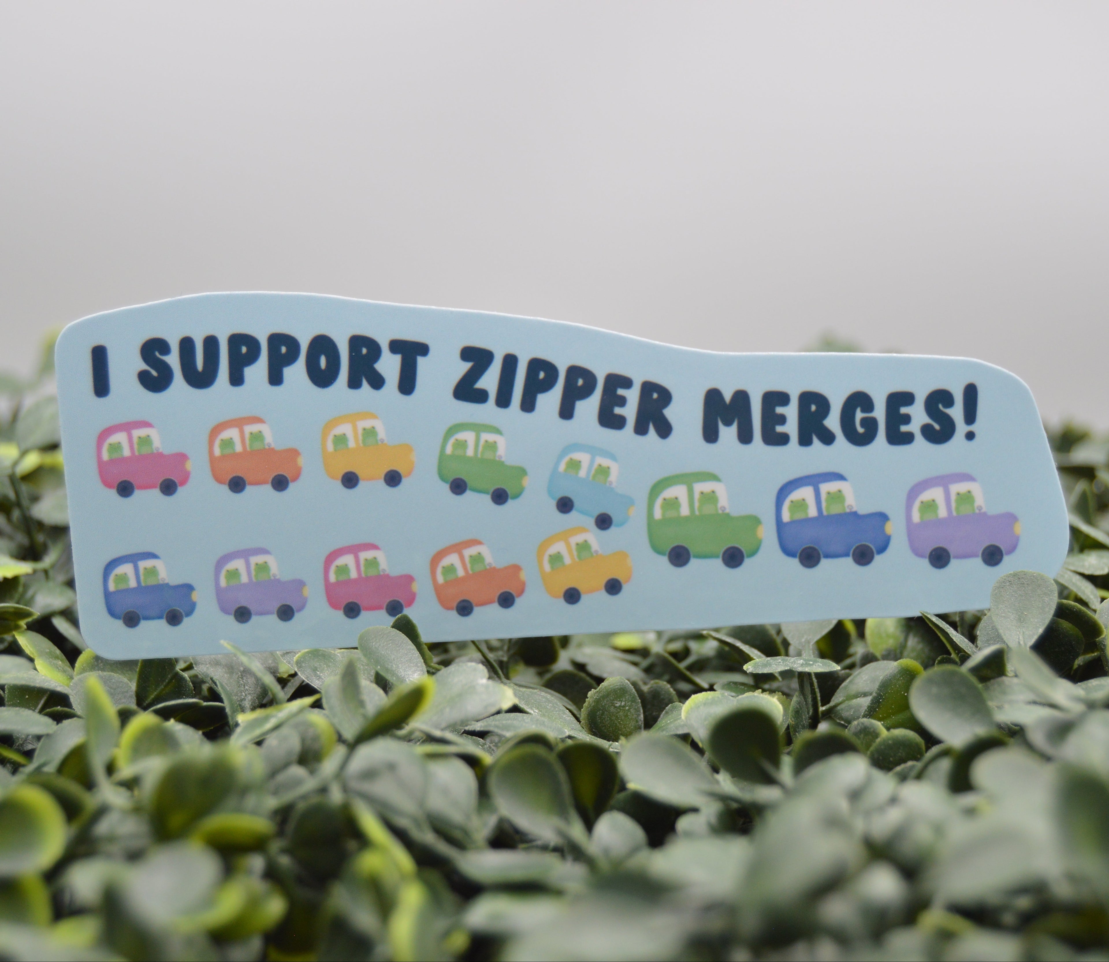 A die-cut sticker with the text 'I SUPPORT ZIPPER MERGES!' and small colorful cartoon cars, shown resting on green foliage against a soft gray background.