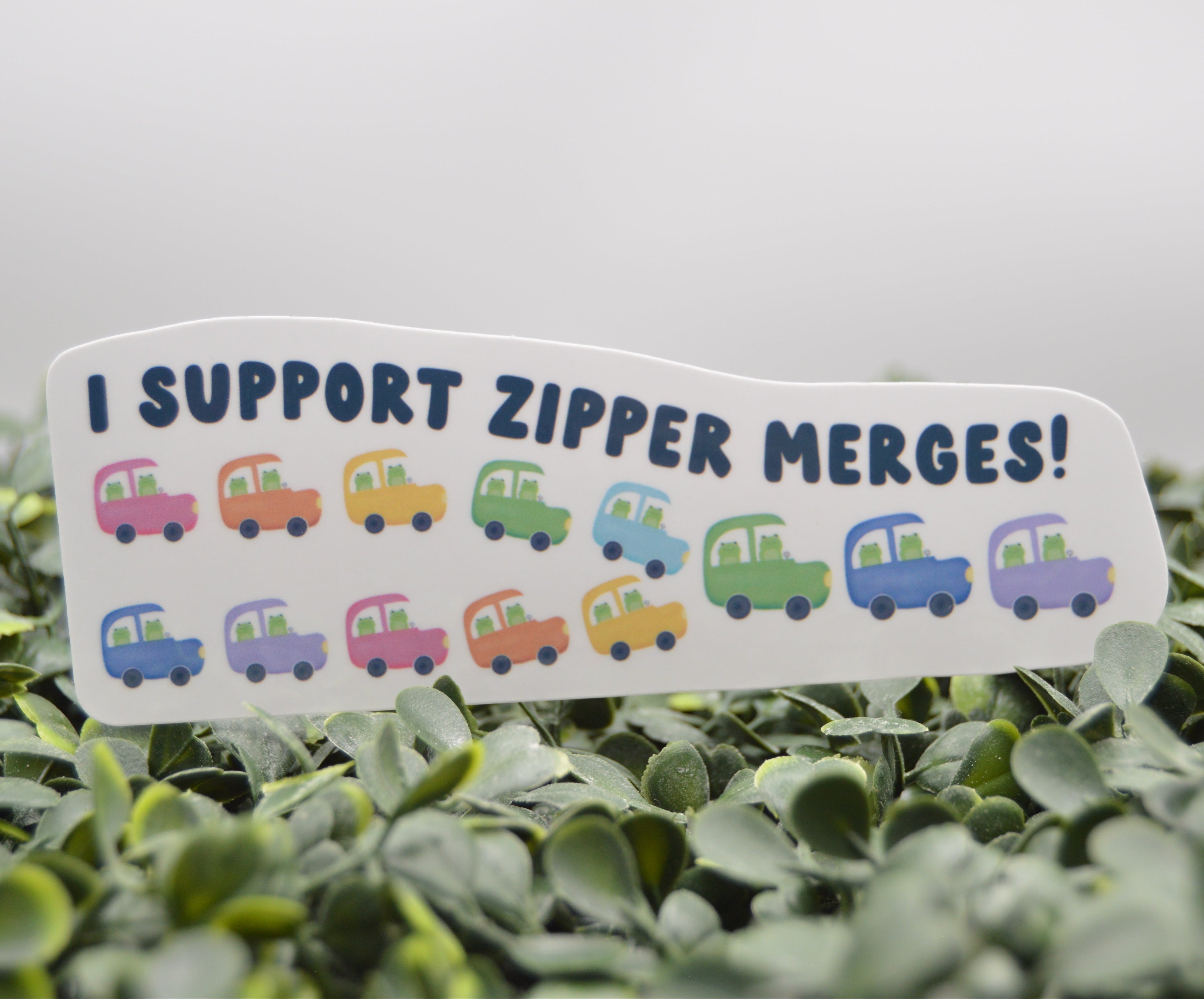 A white die-cut sticker with the text 'I SUPPORT ZIPPER MERGES!' and a row of colorful cartoon cars, displayed in a row of green foliage against a light gray background.