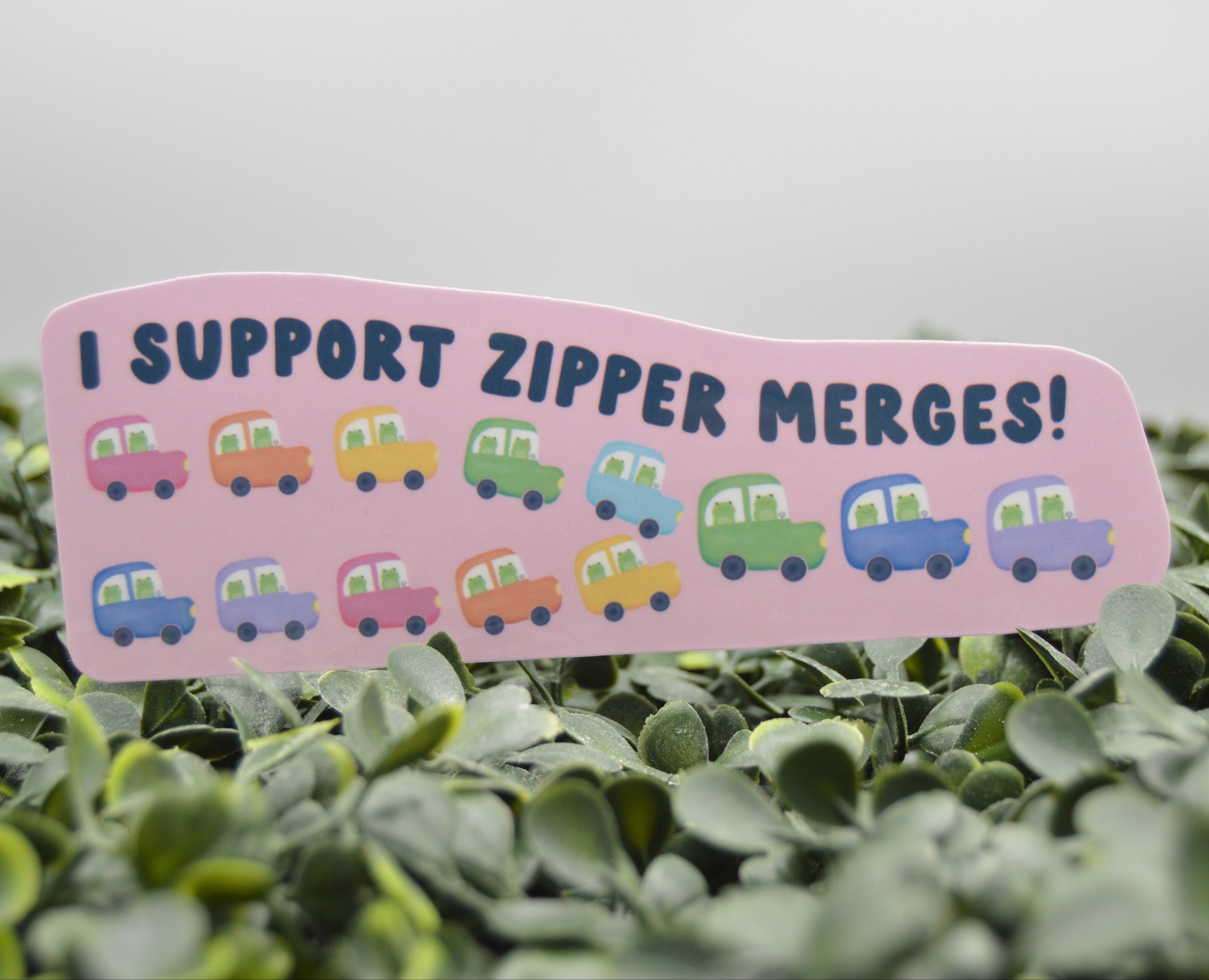 A pink die-cut sticker with the text 'I SUPPORT ZIPPER MERGES!' and small colorful cartoon cars, displayed among green leaves.