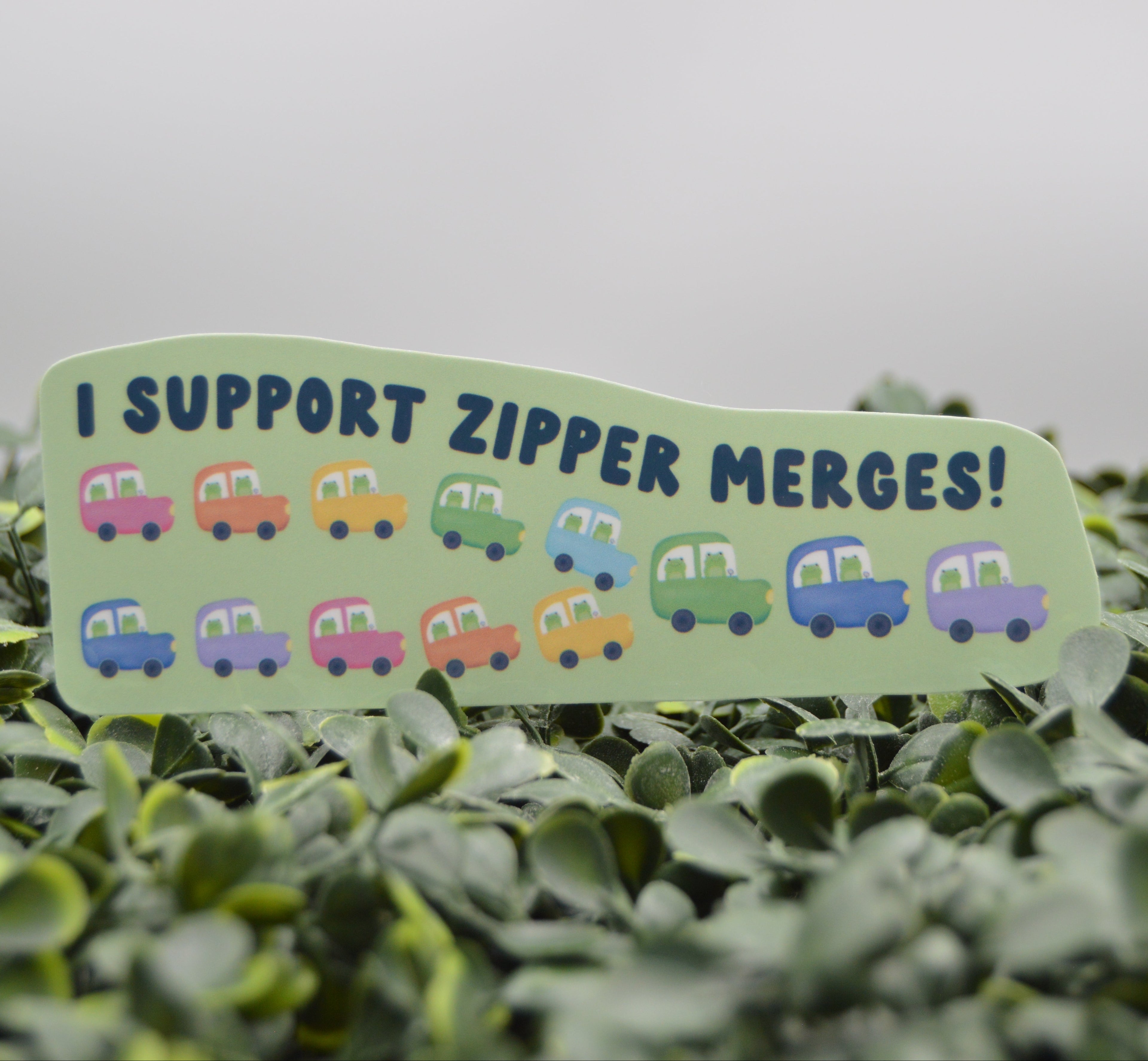 A die-cut sticker with the text 'I SUPPORT ZIPPER MERGES!' and small colorful cartoon cars, placed among green leaves against a soft gray background.