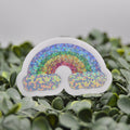 A die-cut sticker shaped like a rainbow with glittery multicolor bands and white cloud-like lower sections, placed on green foliage against a soft gray background.