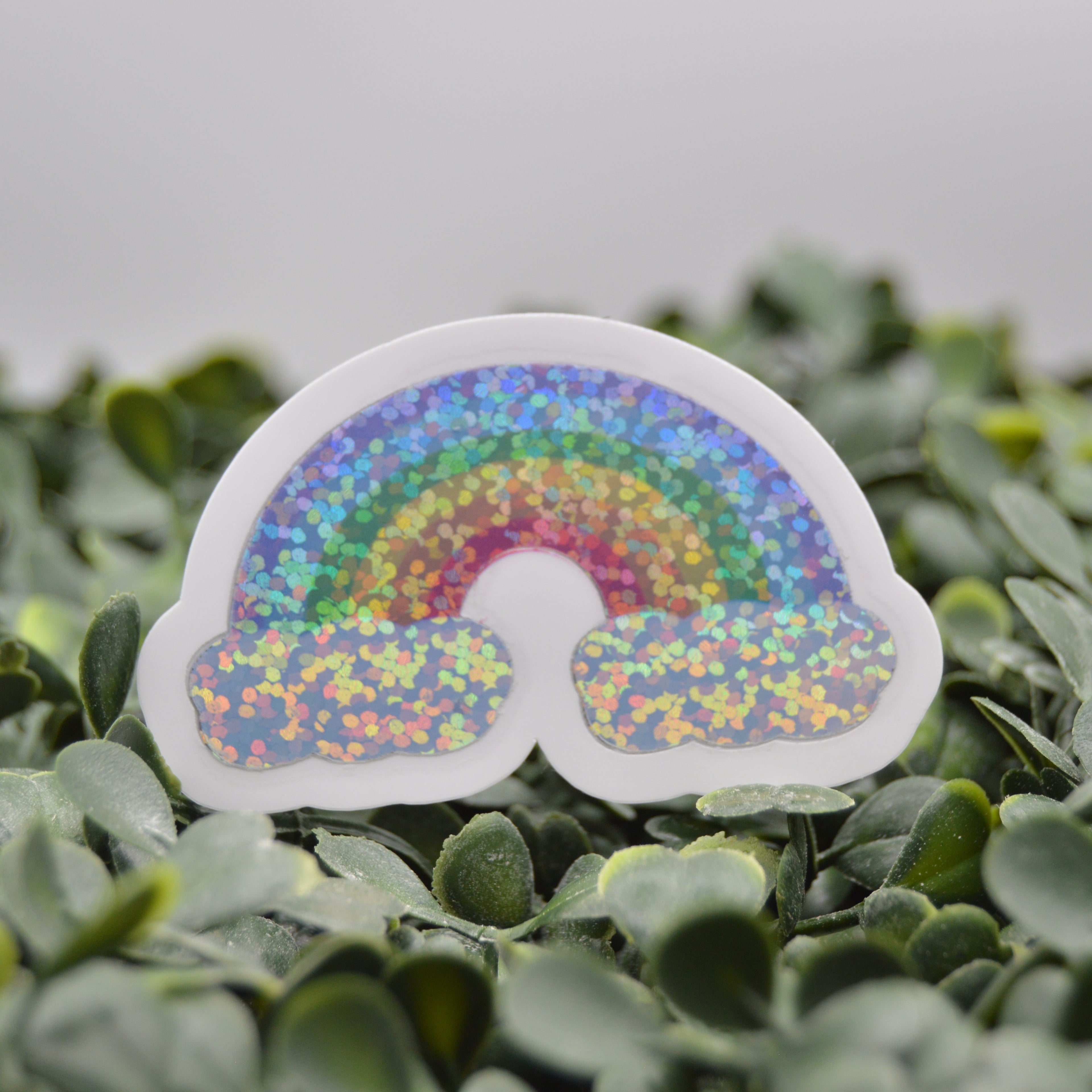 A die-cut sticker shaped like a rainbow with glittery multicolor bands and white cloud-like lower sections, placed on green foliage against a soft gray background.