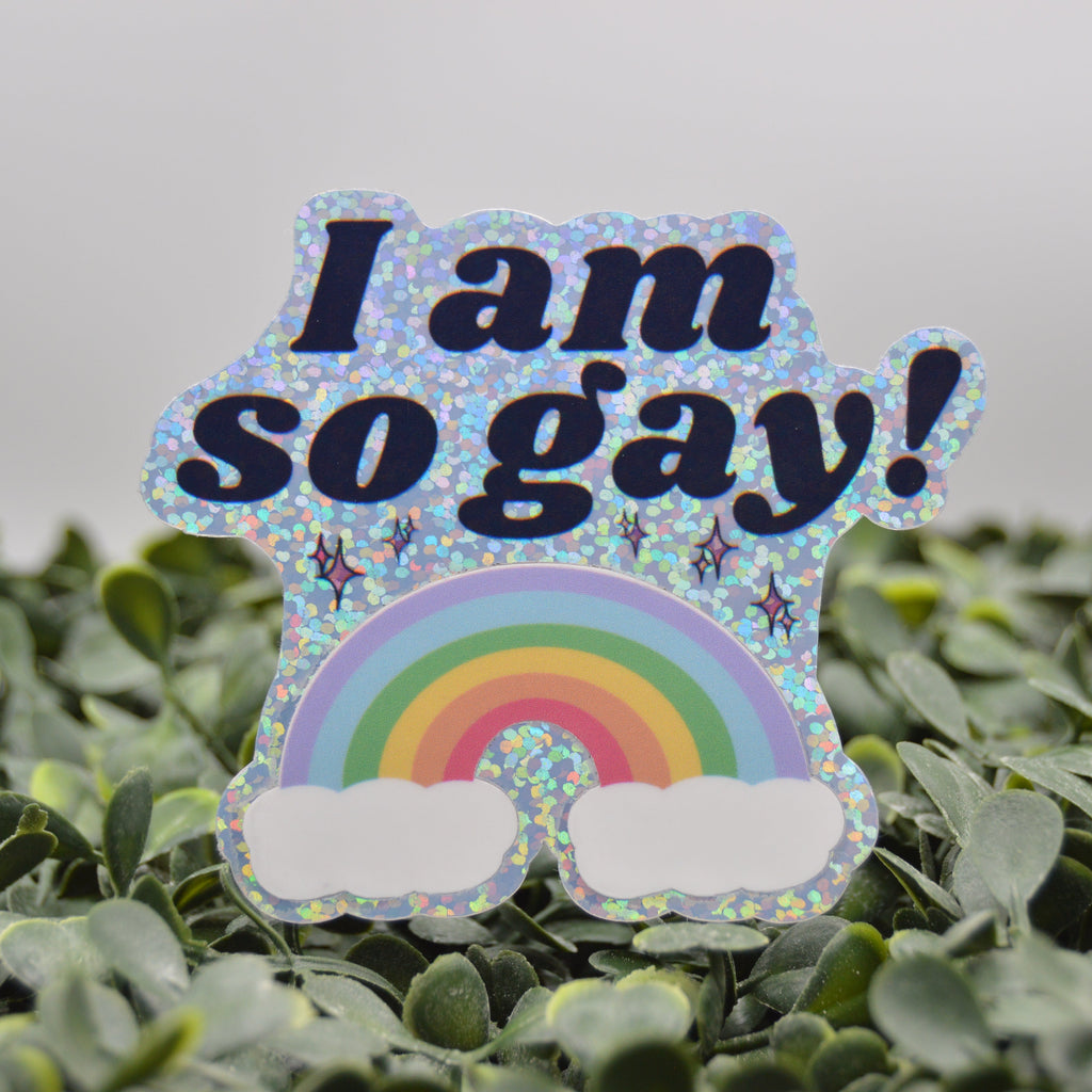 A die-cut sticker with the text 'I am so gay!' in dark blue lettering above a rainbow and white clouds, shown standing upright on green foliage against a soft gray background.