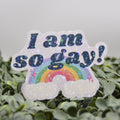 A die-cut sticker with the text 'I am so gay!' in glittery dark blue lettering above a rainbow and clouds, displayed on green foliage against a soft gray background.