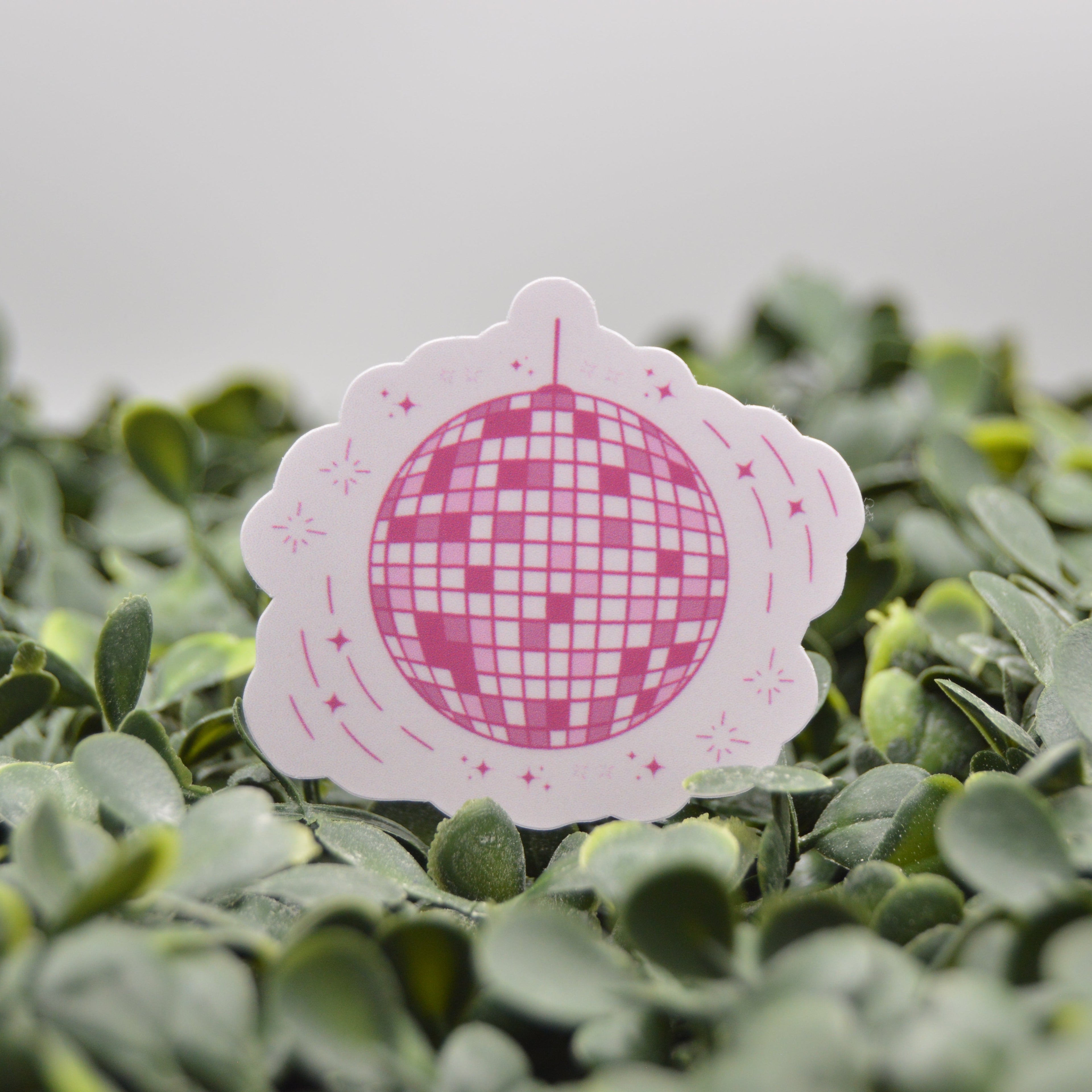 A die-cut sticker featuring a pink and white disco ball design on a scalloped white border, displayed on a bed of green leaves against a soft gray background.