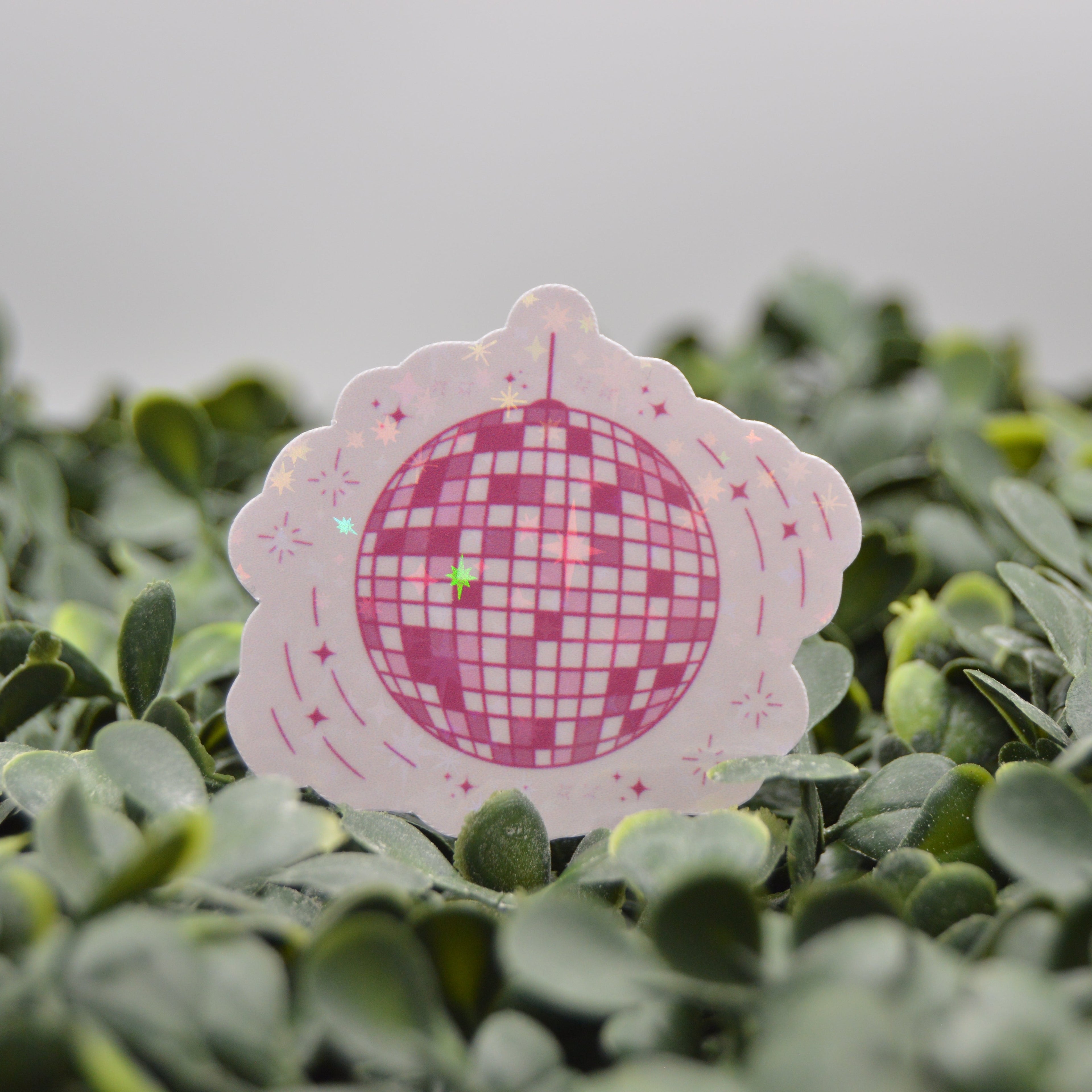 A pink and white scalloped die-cut sticker featuring a disco ball design, placed on a bed of green leaves.