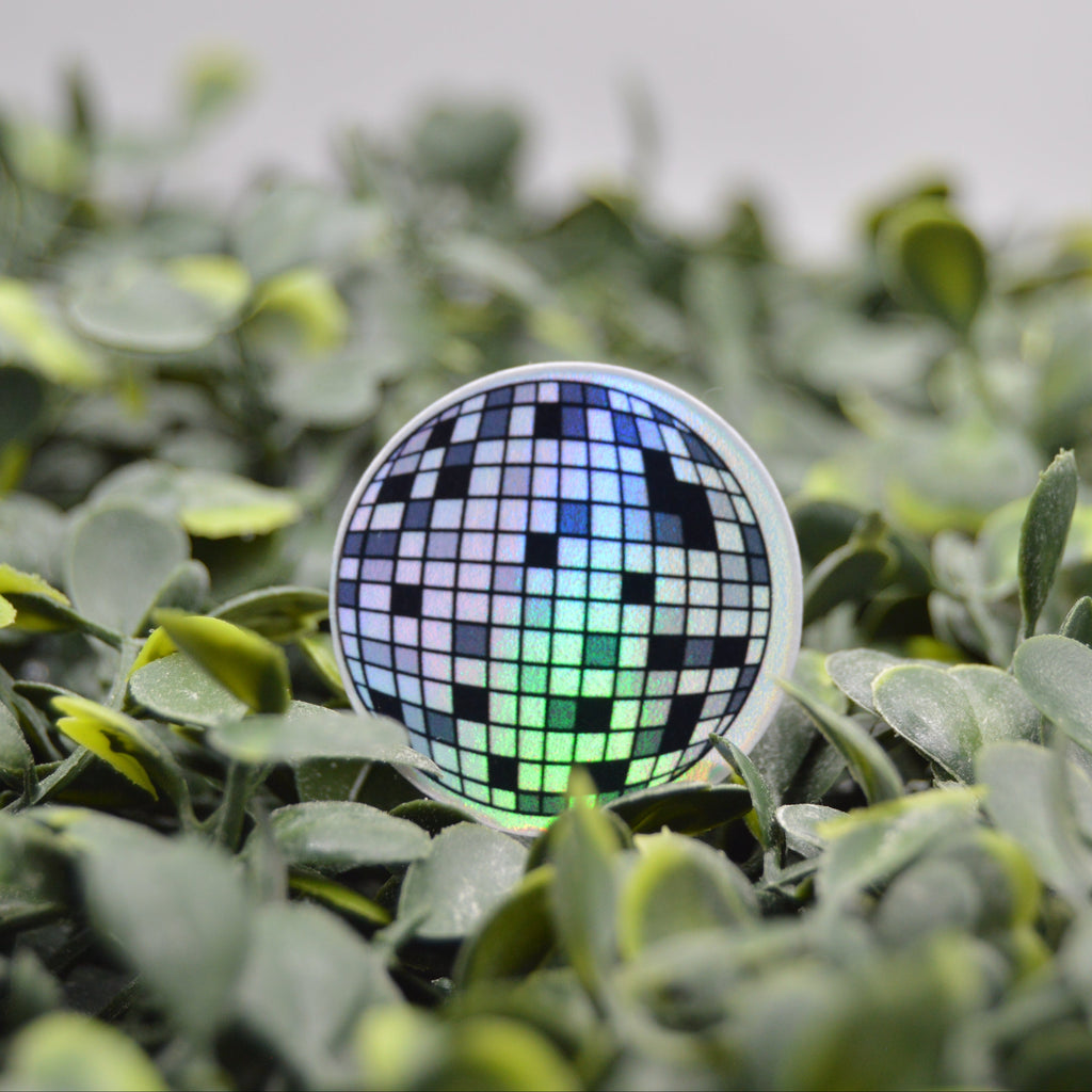 A round sticker or patch featuring a disco ball design in blue, green, and black, resting on a bed of green leaves.