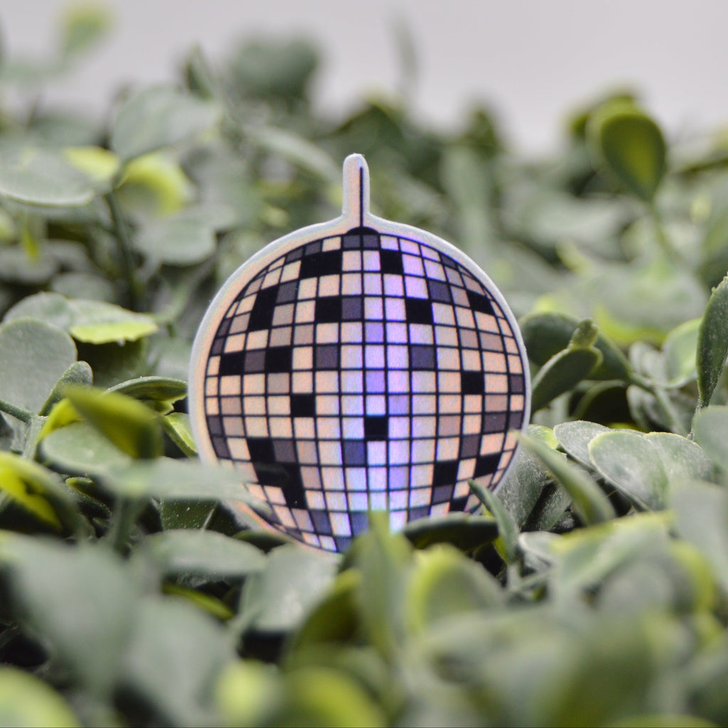 A small disco ball ornament with a black, white, and purple pixelated pattern, nestled among green leaves.