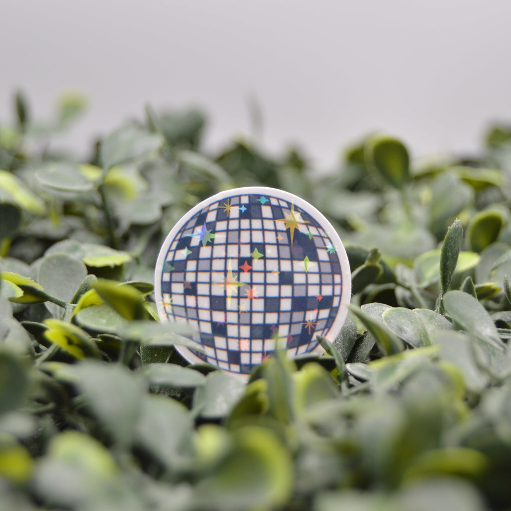 A round pinback button badge featuring a blue and white checkered disco ball pattern with colorful star accents, displayed on a bed of green artificial foliage.