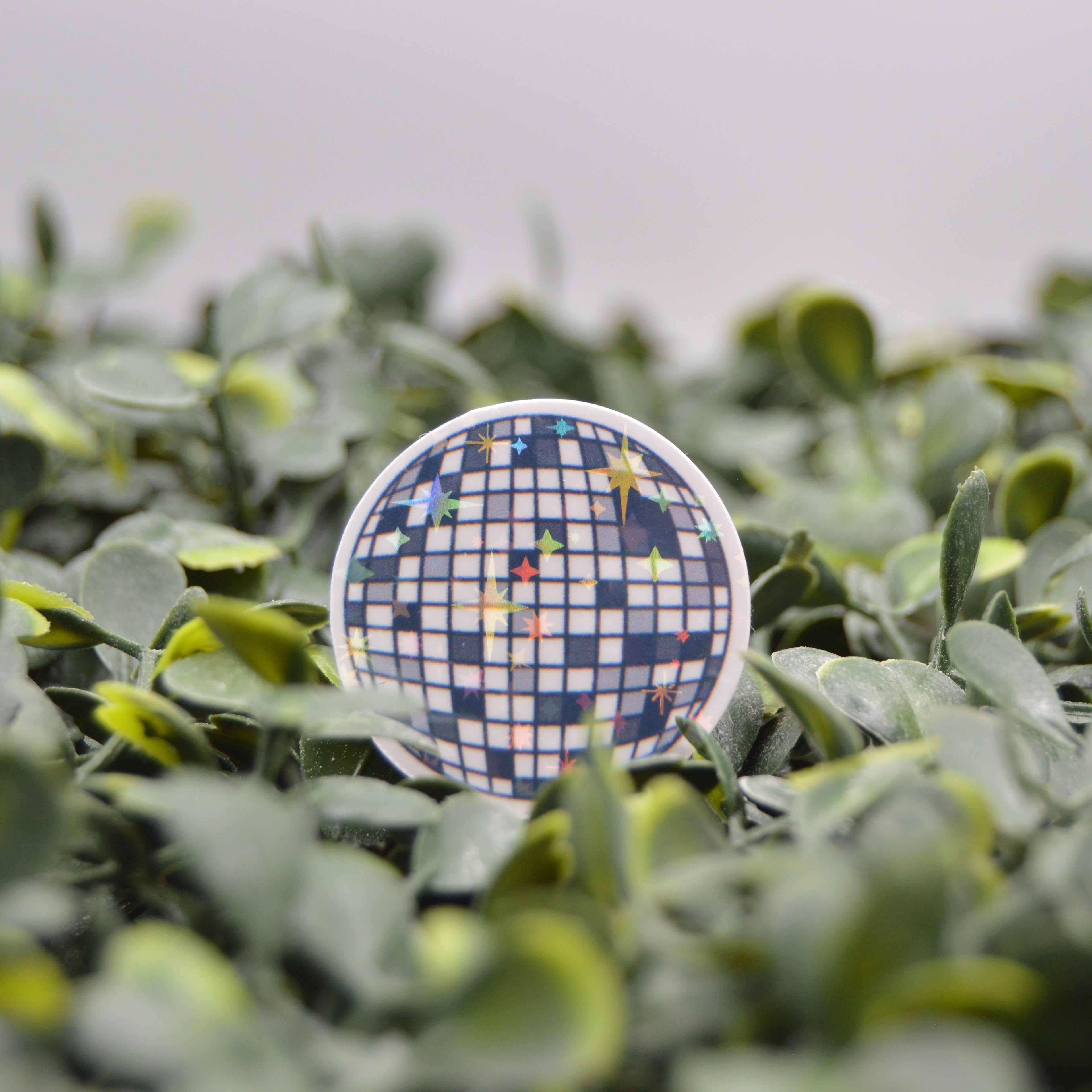 A round pinback button badge featuring a blue and white checkered disco ball pattern with colorful star accents, displayed on a bed of green artificial foliage.
