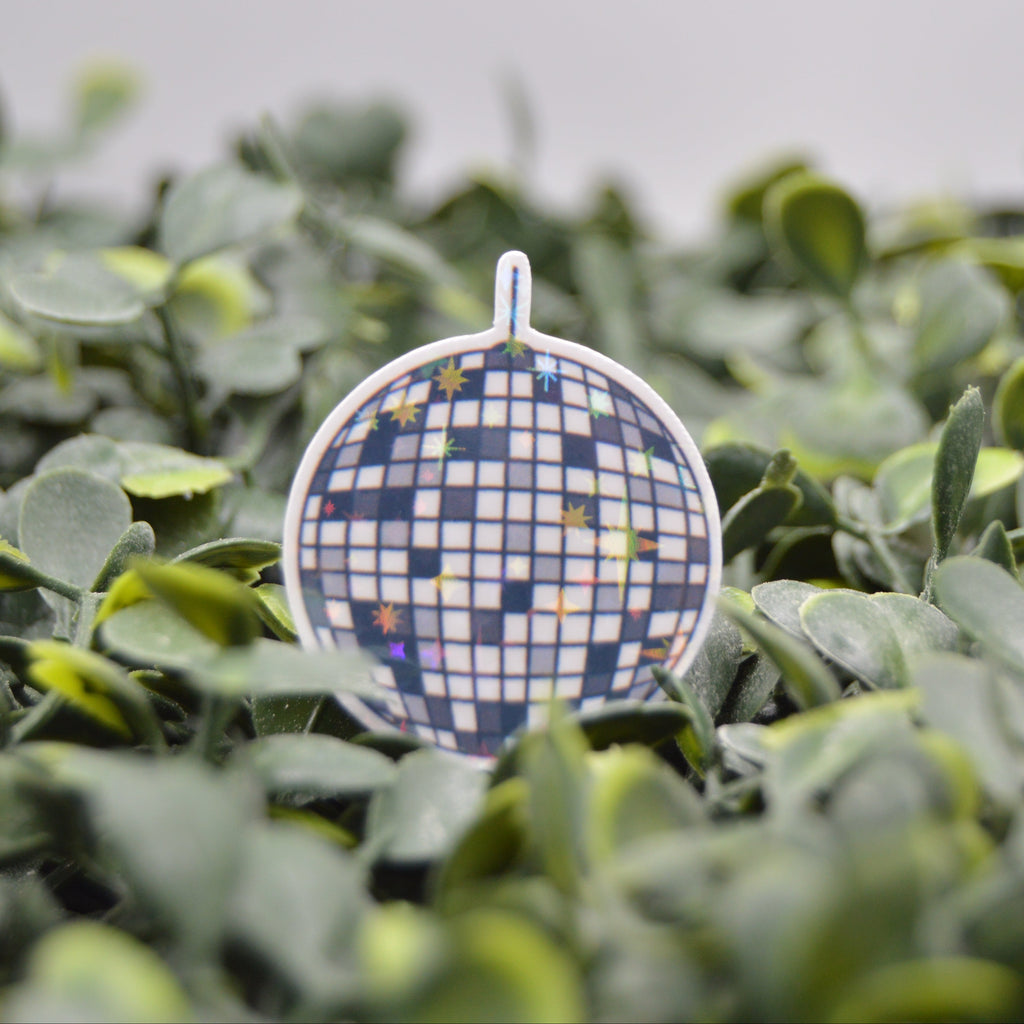 A small round ornament or charm with a blue, white, and yellow mosaic disco-ball pattern, standing upright amidst green leaves.