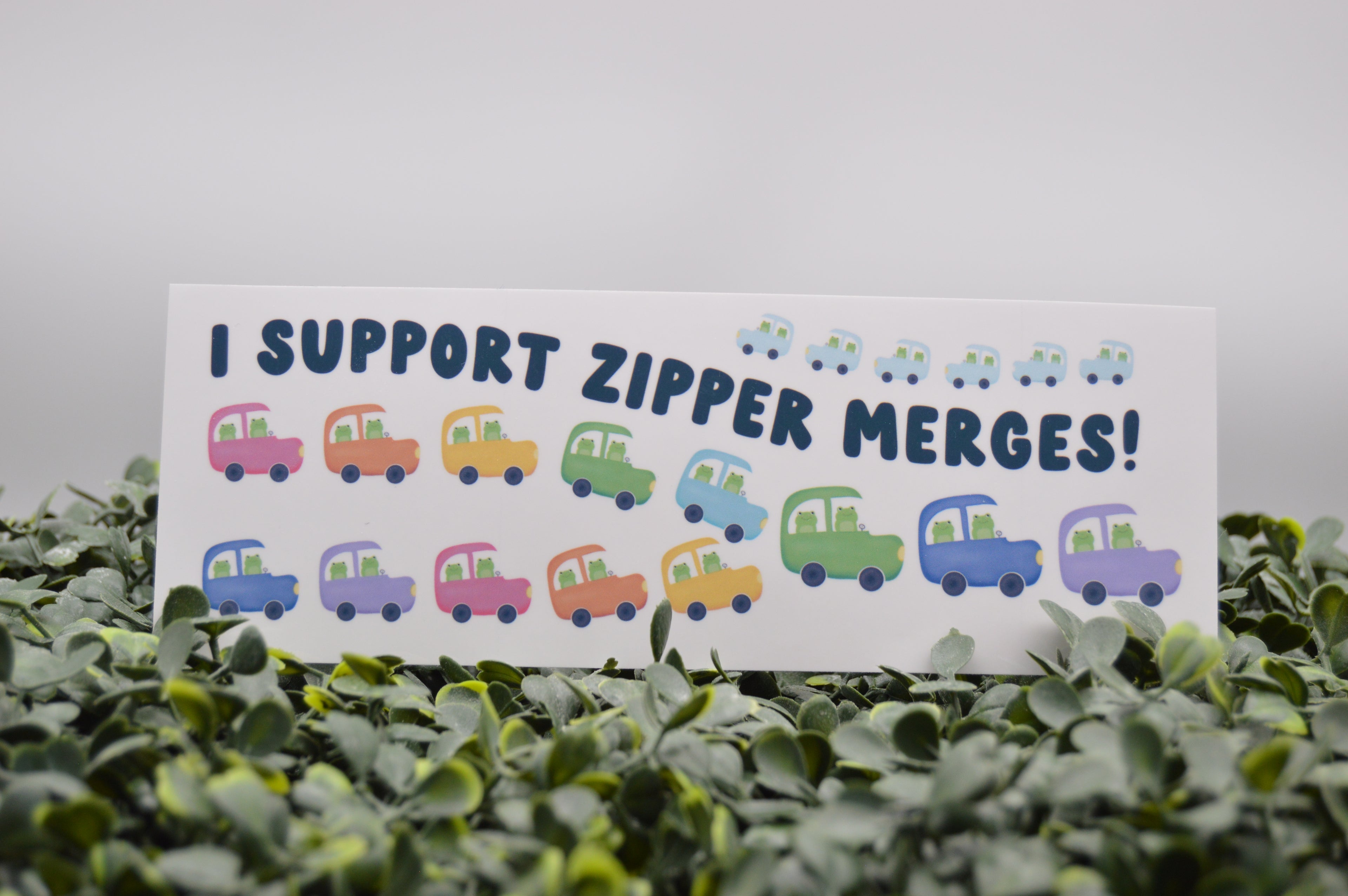 A rectangular white sign with the text 'I SUPPORT ZIPPER MERGES!' in dark blue, surrounded by small colorful cartoon cars, placed among green leaves against a soft gray background.