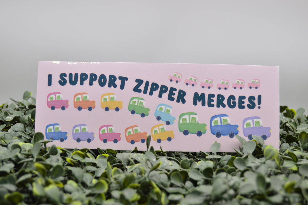 A rectangular pink sign with the text 'I SUPPORT ZIPPER MERGES!' and rows of small colorful cartoon cars, placed among green leaves against a soft gray background.