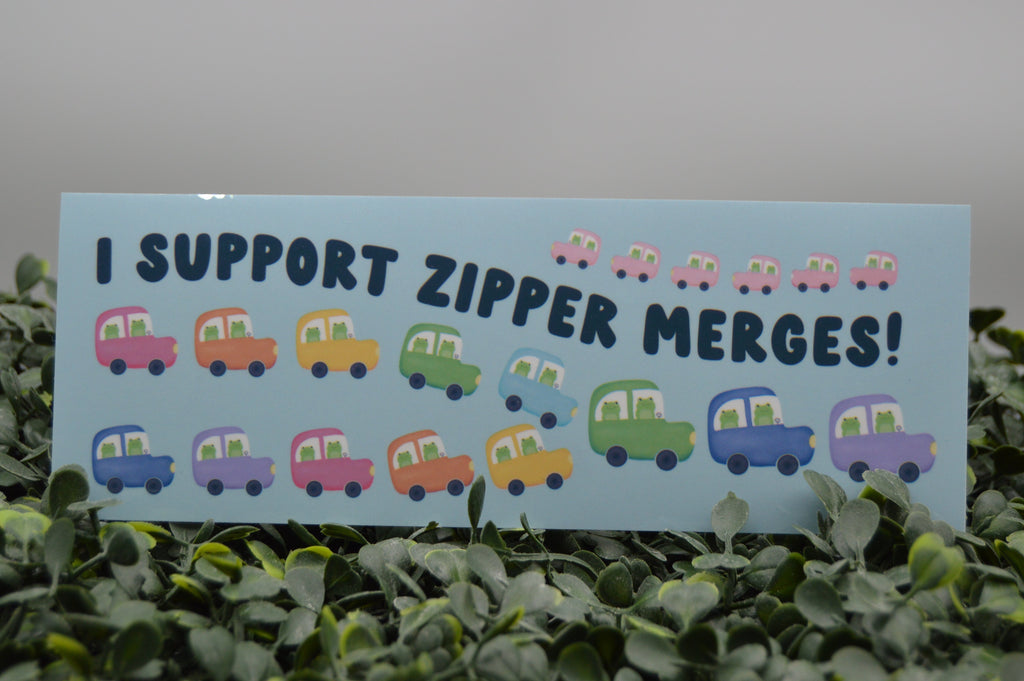 A rectangular light blue sign with the text 'I SUPPORT ZIPPER MERGES!' in dark blue letters, decorated with cartoon cars in pink, yellow, green, blue, and purple, displayed among green leaves against a gray background.