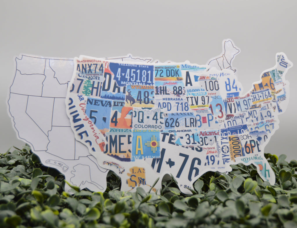 A white silhouette map of the United States filled with colorful vintage license plates, displayed on a bed of green foliage against a light gray background.