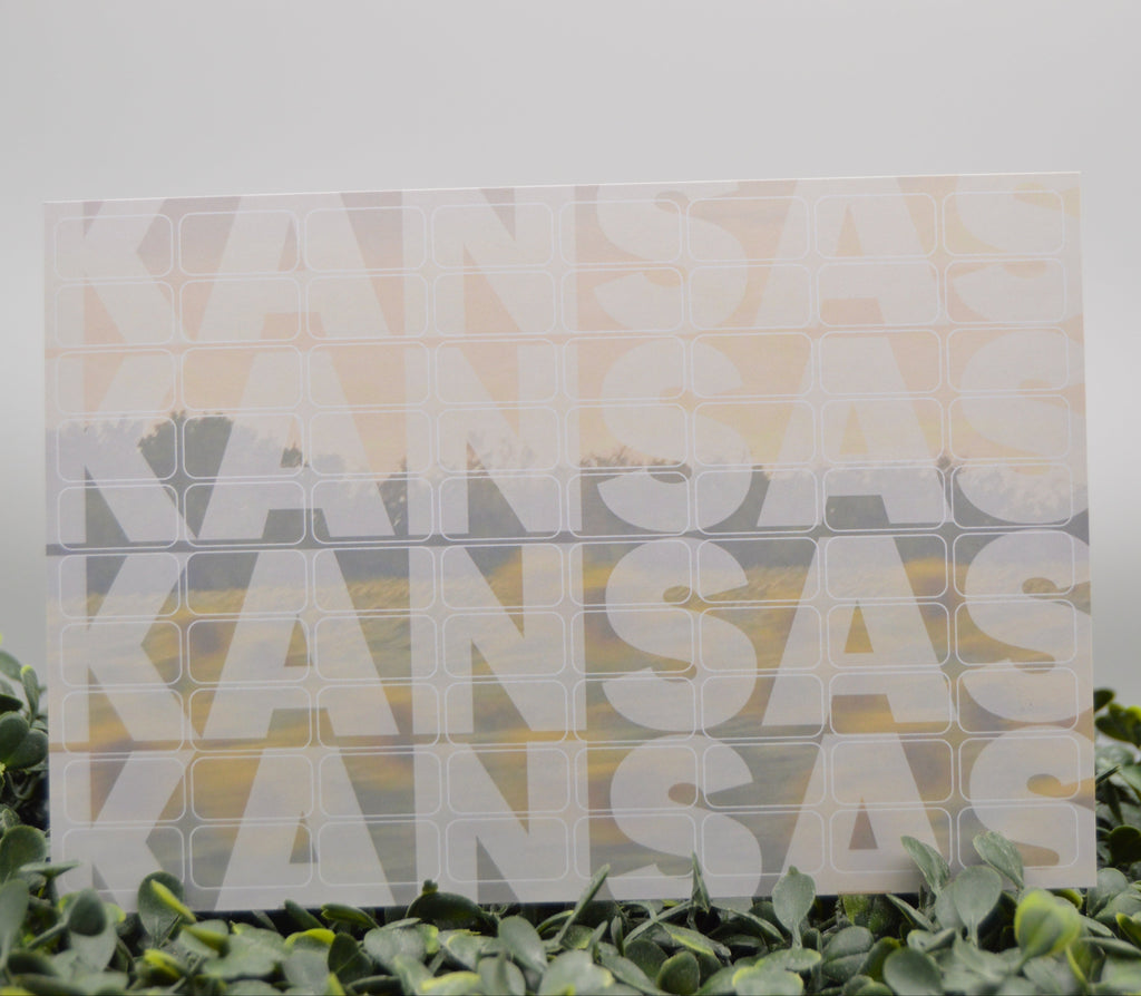 A square acrylic art print featuring the word 'KANSAS' repeated in large white letters over a soft landscape gradient of yellow, orange, and gray. The print is styled standing upright on a bed of green eucalyptus leaves against a neutral light gray background.